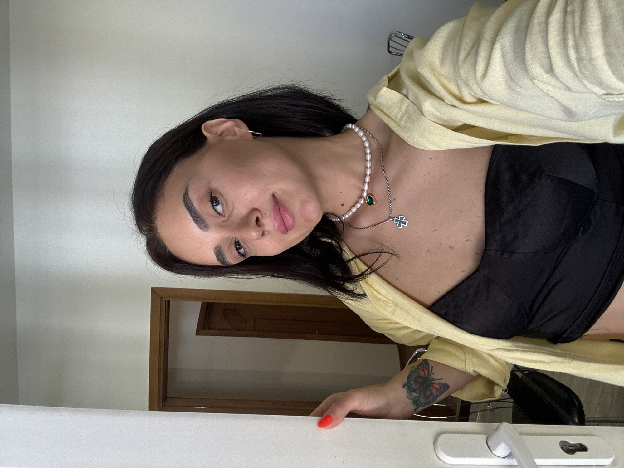 A woman with short, dark hair and fair skin stands in a doorway, looking at the camera. She is wearing a black strapless top and a light yellow open button-up shirt over it. She has a pearl necklace with a green pendant and a silver chain with a blue cross pendant. A butterfly tattoo is visible on her left arm. Her fingernails are painted bright red. To her left, a white door is partially open, revealing a white door handle and lock mechanism. In the background, there is a hallway with wooden doors. The lighting is bright and appears to be natural light coming from the right. The image is taken from the chest up. The location is identified as Tbilisi, Georgia.