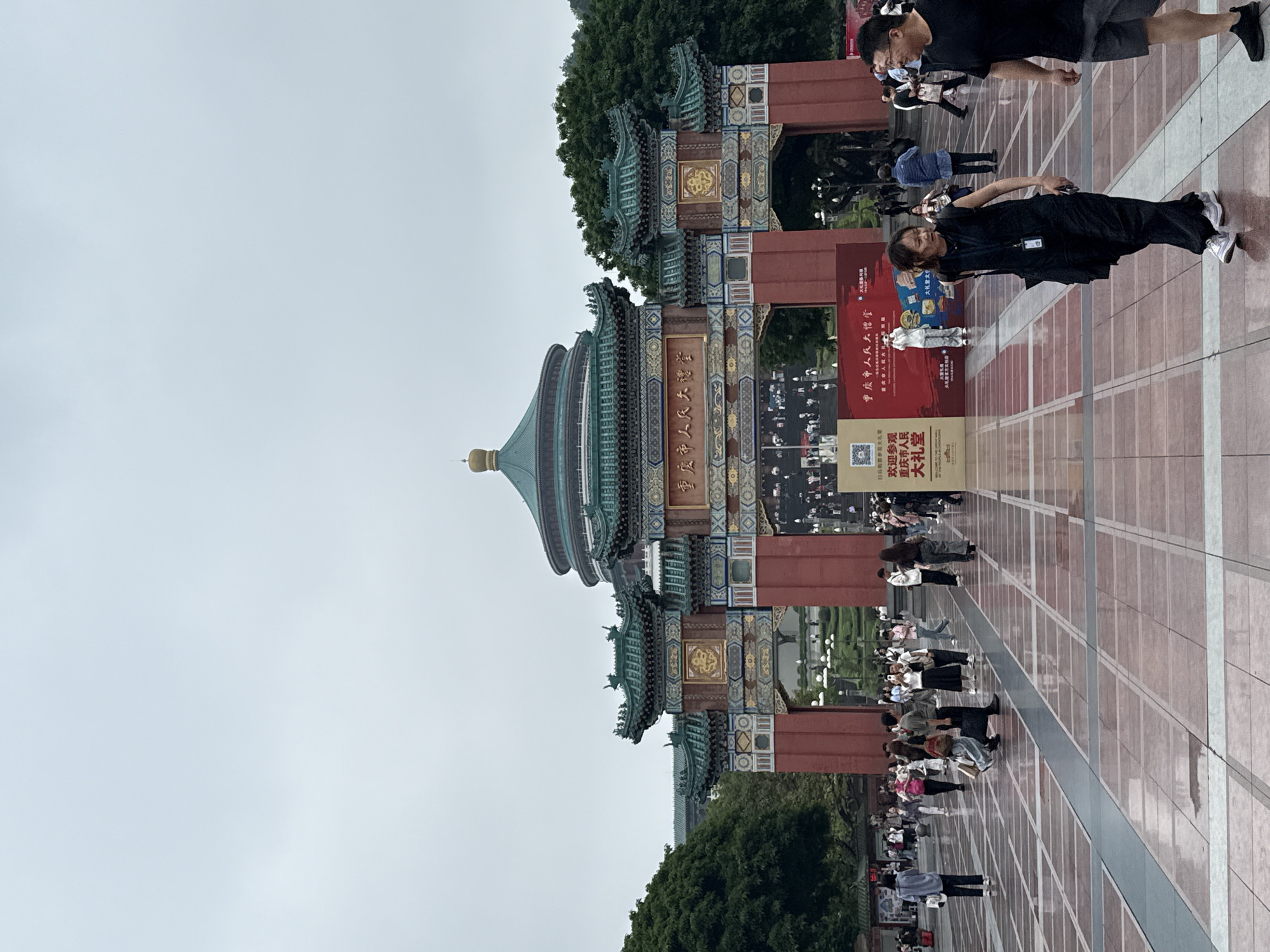 The image depicts Chongqing People's Hall in Yuzhong District, China, featuring its distinctive traditional Chinese architecture. People are milling about in the open plaza. Signage near the building is prominently displayed with Chinese characters. The sky appears overcast.