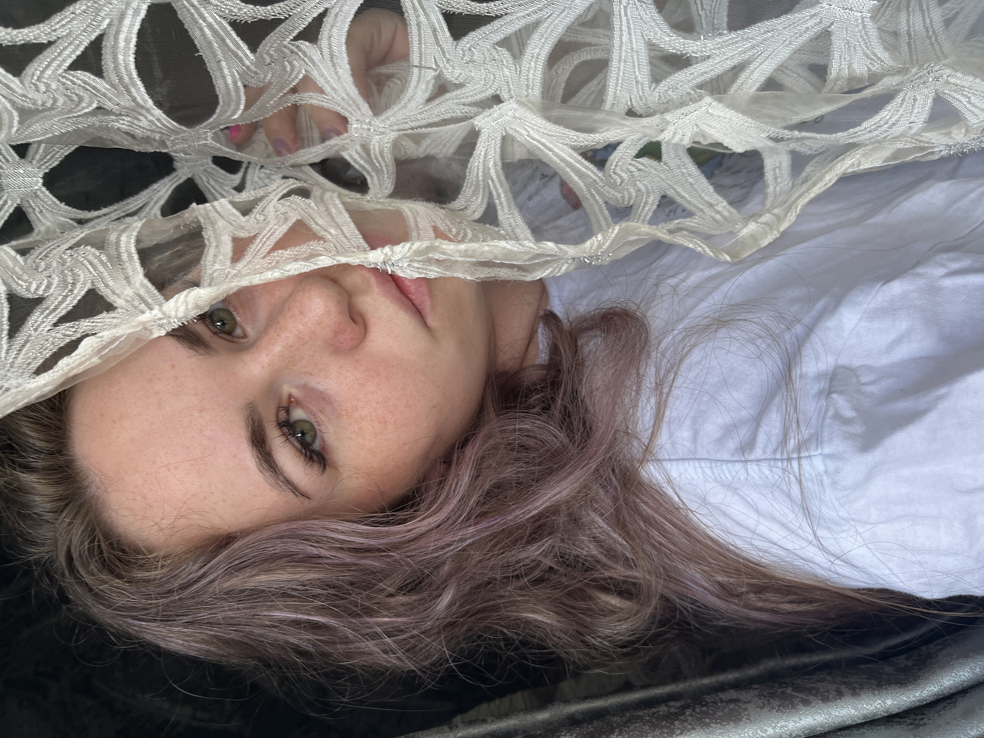 This media file presents a close-up, horizontally oriented portrait of a young individual, likely female, captured from an overhead perspective. The subject has fair skin with visible freckles across the nose and cheeks, and striking green eyes with dark eyelashes. Her light brown hair features subtle purple or pinkish undertones and is styled in soft waves, fanning out around her head.
A significant portion of the upper face and head is partially obscured by an intricate, off-white or cream-colored sheer fabric, possibly lace or embroidered netting, featuring an open-work, geometric pattern. A hand with bright pink/purple painted fingernails is visible through this sheer fabric in the upper left of the frame, appearing to hold or adjust the material. The subject's mouth is subtly parted.
She appears to be lying down, dressed in a white fabric, possibly a shirt or bedsheet, which is visible beneath her hair and shoulder. Below this, a dark, possibly metallic or reflective surface can be seen. The lighting is soft and diffused, characteristic of an indoor setting, and does not indicate a specific time of day. The subject's expression is neutral and direct, gazing towards the camera. No text is visible in the image. The scene takes place in Chaikovsky, Russia.