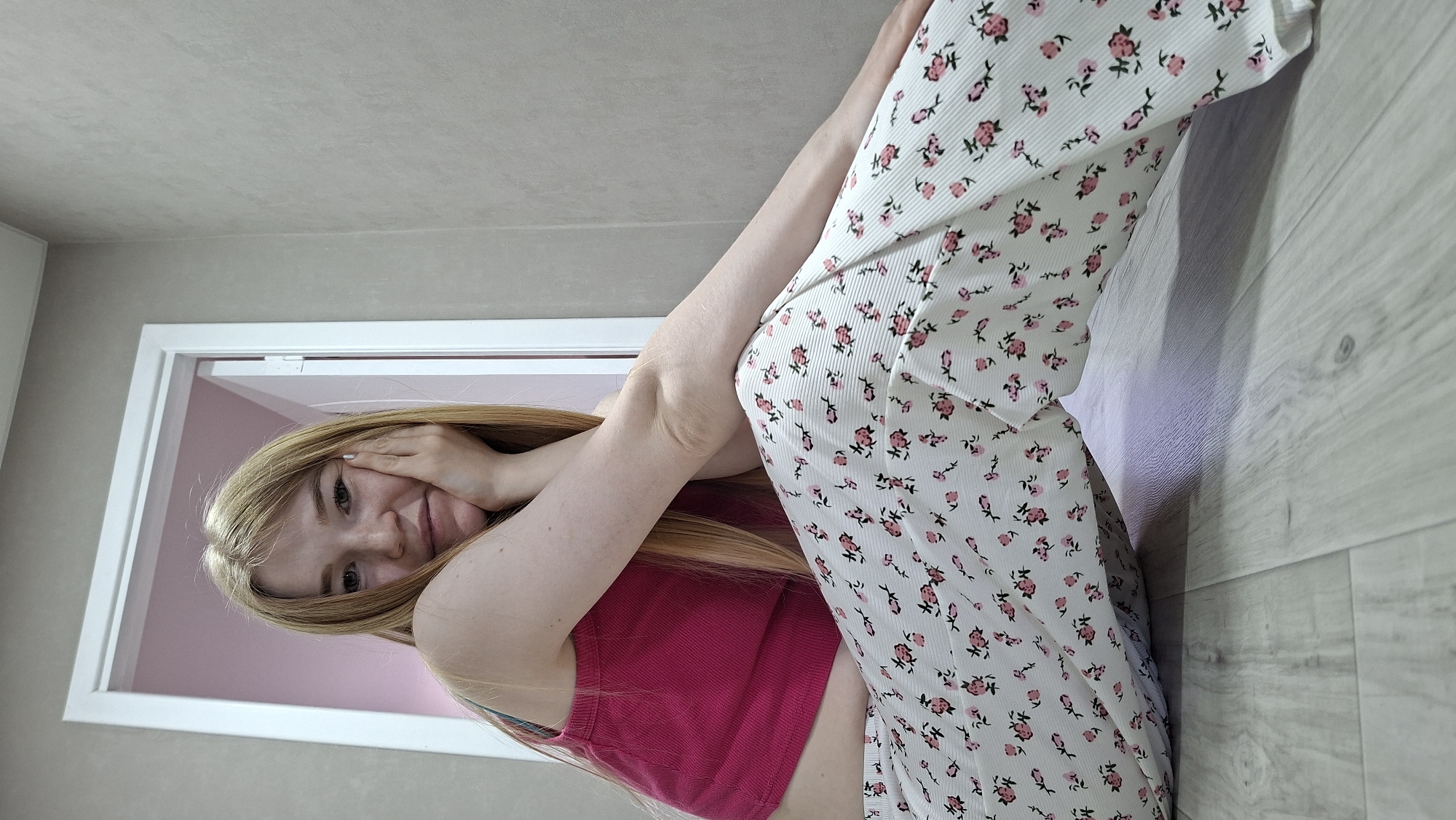 This is an indoor portrait of a young woman with long, straight blonde hair, located in Chaikovsky, Russia. The image is taken from a low angle and is rotated, depicting the subject lying on a light wood-patterned floor, with a light grey wall extending upwards from her.
The woman is wearing a bright fuchsia (pink) sleeveless top and white pants patterned with small pink flowers and green stems/leaves. Her body is positioned diagonally across the frame, with her legs extended towards the upper right. She is looking directly at the camera with a soft, friendly smile. One of her hands is gently placed on the side of her face, partially obscuring it, which conveys a sense of shyness or playfulness.
In the background, a white doorframe is visible, opening into another room that has light pink walls. The overall lighting in the scene is bright and even, suggesting an indoor setting during the daytime. The environment appears to be a relaxed home interior. There is no visible text in the image.