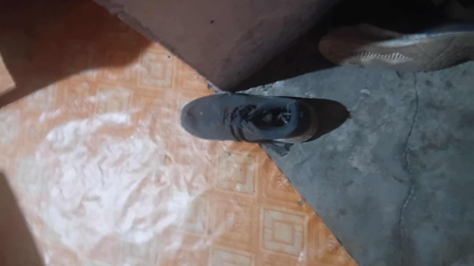 In the city of Prabumulih, Indonesia, a dark-colored sneaker is placed on a cracked concrete surface adjacent to a tiled floor. The tiled floor has a pattern of square tiles in a light brown hue with darker brown square outlines. The sneaker is positioned with its toe pointing towards the upper left of the frame. To the right of the sneaker, a large, dark object, possibly a box or furniture, casts a shadow. The lighting in the scene is dim, with some areas illuminated by a light source that creates bright, diffused reflections on the tiled floor. The overall environment suggests an indoor or covered outdoor space with utilitarian flooring.