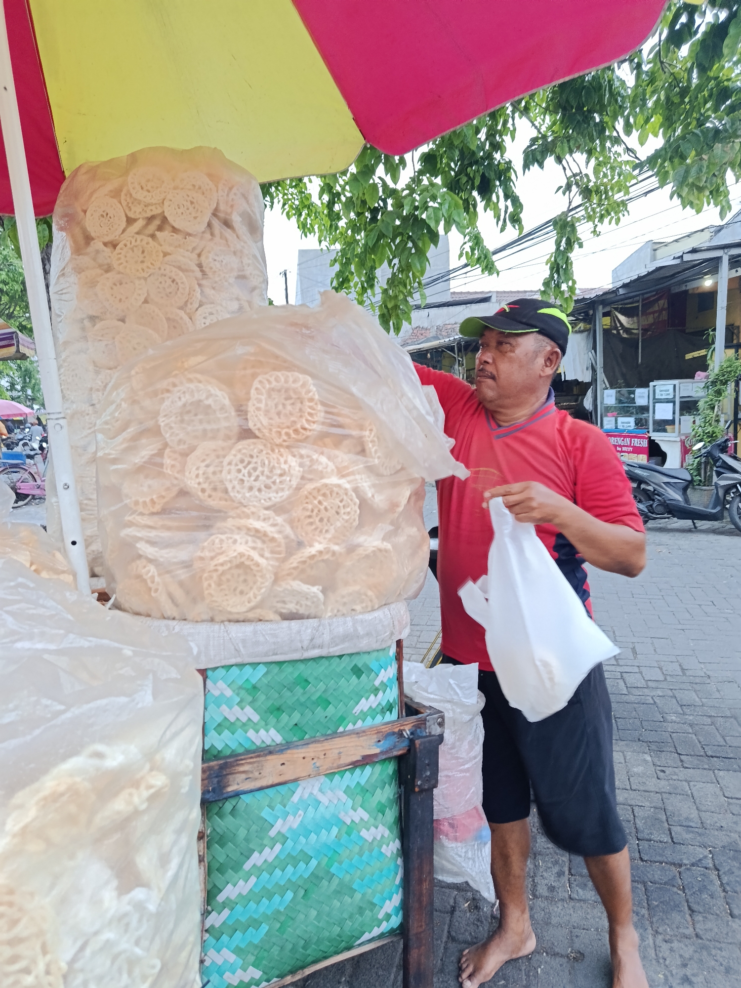 A middle-aged man wearing a red t-shirt and black shorts stands barefoot on a paved sidewalk in Keboansikep, Indonesia, next to a street food stall. He is holding a white plastic bag in his right hand and is reaching into a large plastic bag filled with circular, white, lace-like crackers. The stall is shaded by a yellow and pink umbrella. Behind the man, there are buildings with open fronts, and a motorcycle is parked on the sidewalk. The man's left hand is near the top of the cracker bag. The pavement is made of grey interlocking bricks.