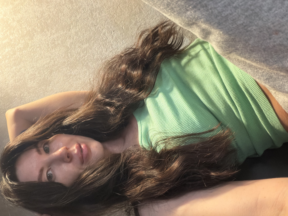 A single female subject is depicted in a medium-close up, reclining indoors. The individual has long, wavy, dark brown hair, parted on the left, cascading over her shoulders and chest. Her face, oriented towards the camera, exhibits fair skin, greenish-grey eyes, and naturally colored, slightly parted lips. Her right arm is raised and bent, with the hand positioned behind her head or neck. She is wearing a ribbed, light mint green tank top, and light grey casual trousers are partially visible on her left leg. Direct light from the left casts parallel striped shadows across her upper torso and the textured, light-colored wall in the background. The scene is located in Perm, Russia.