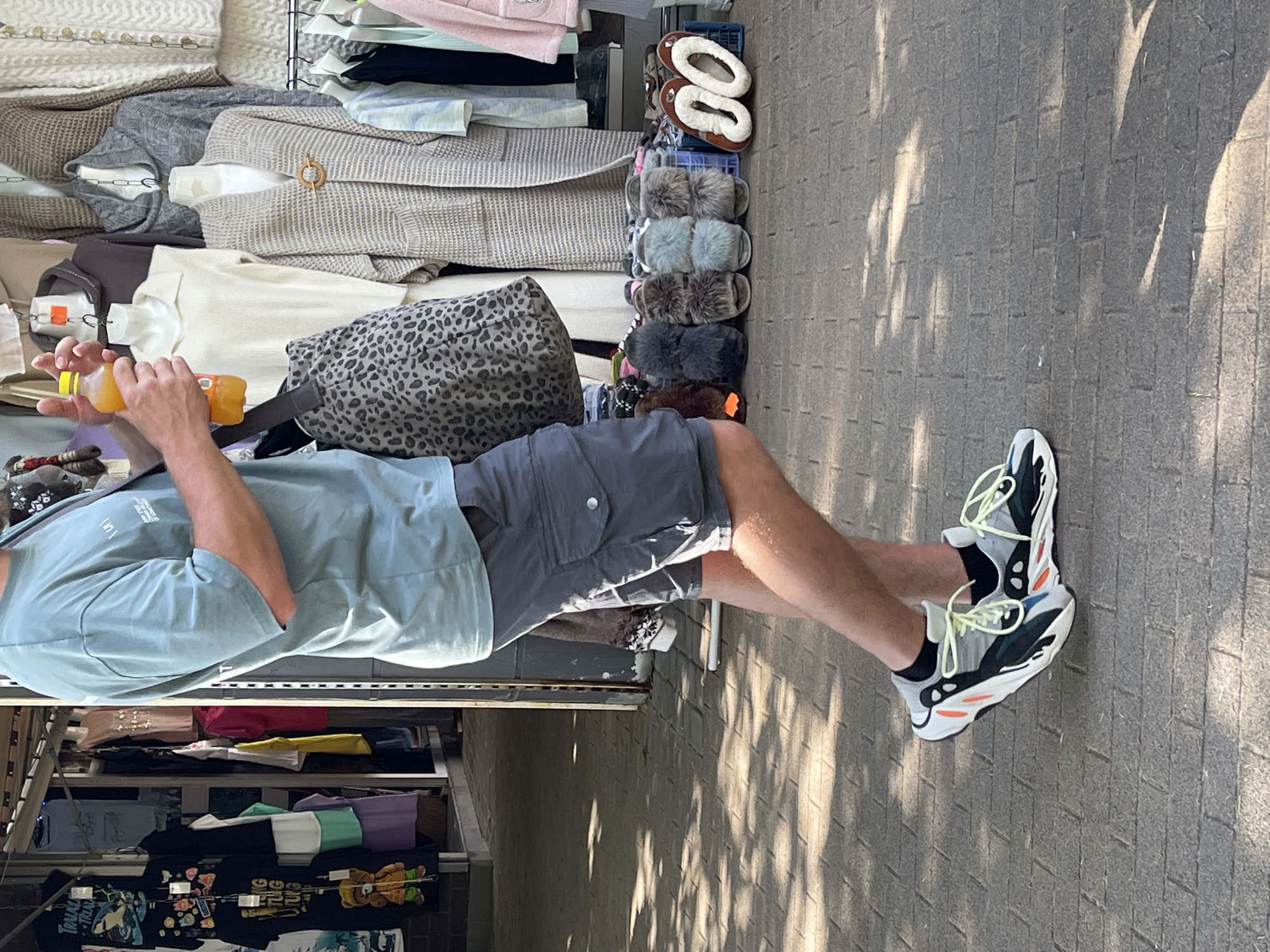 A person is standing on a brick sidewalk in front of a clothing stall. They are wearing a teal t-shirt, grey cargo shorts, and athletic shoes. In their right hand, they are holding a yellow bottle of liquid. Behind the person, clothing is displayed on mannequins and racks. Several pairs of fuzzy slippers and other footwear are arranged on the ground in front of the stall. The ground is partially covered by dappled sunlight and shadows.