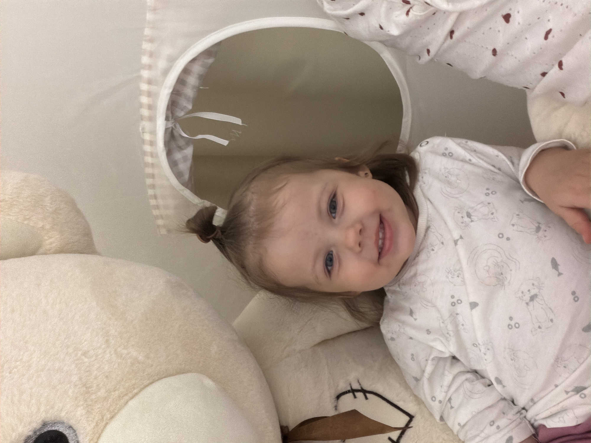 A young child with blue eyes and a small ponytail is lying on a plush, cream-colored teddy bear. She is wearing a white long-sleeved shirt with a pattern of animals. The child is smiling and appears to be looking up at the camera. Above her is a white fabric tunnel or play structure with a circular opening, suggesting she might be in a play area. The scene appears to be indoors. There is no verifiable information to suggest this image was taken in the city of Reshetnikovo, Russia. The location information is not possible to determine from the image.