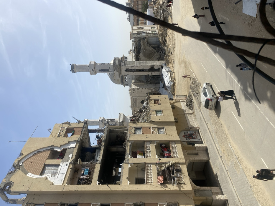 The image depicts a devastated urban landscape in Khan Yunis, Palestinian Territory, under a clear, bright blue sky with faint cirrus clouds. The scene is dominated by a multi-story residential building on the left, exhibiting extensive structural damage, including collapsed walls, exposed interiors across multiple floors, and shattered windows. Balconies on the lower levels of this building still feature laundry hanging. Impact marks are visible on the facade.
Across a paved street with a visible white dividing line, a heavily damaged mosque stands in the mid-ground. Its white minaret, though showing damage, remains largely intact and upright, while the main mosque structure behind it features a dome and significant wall collapses, exposing its interior.
The ground comprises a mix of dusty, rubble-strewn unpaved areas and a paved road. Piles of debris are scattered throughout. In the unpaved area adjacent to the mosque, white tarpaulins or makeshift shelters are visible among more rubble.
Several individuals are present: two adults converse on the street next to a silver hatchback car. Further down the street, two individuals walk away from the camera. A person in a red shirt walks in the dusty area near the mosque. Additional indistinct figures are visible in the background, walking near the mosque. The photograph appears to be taken from an elevated position, with a blurred metal railing in the immediate foreground on the right.