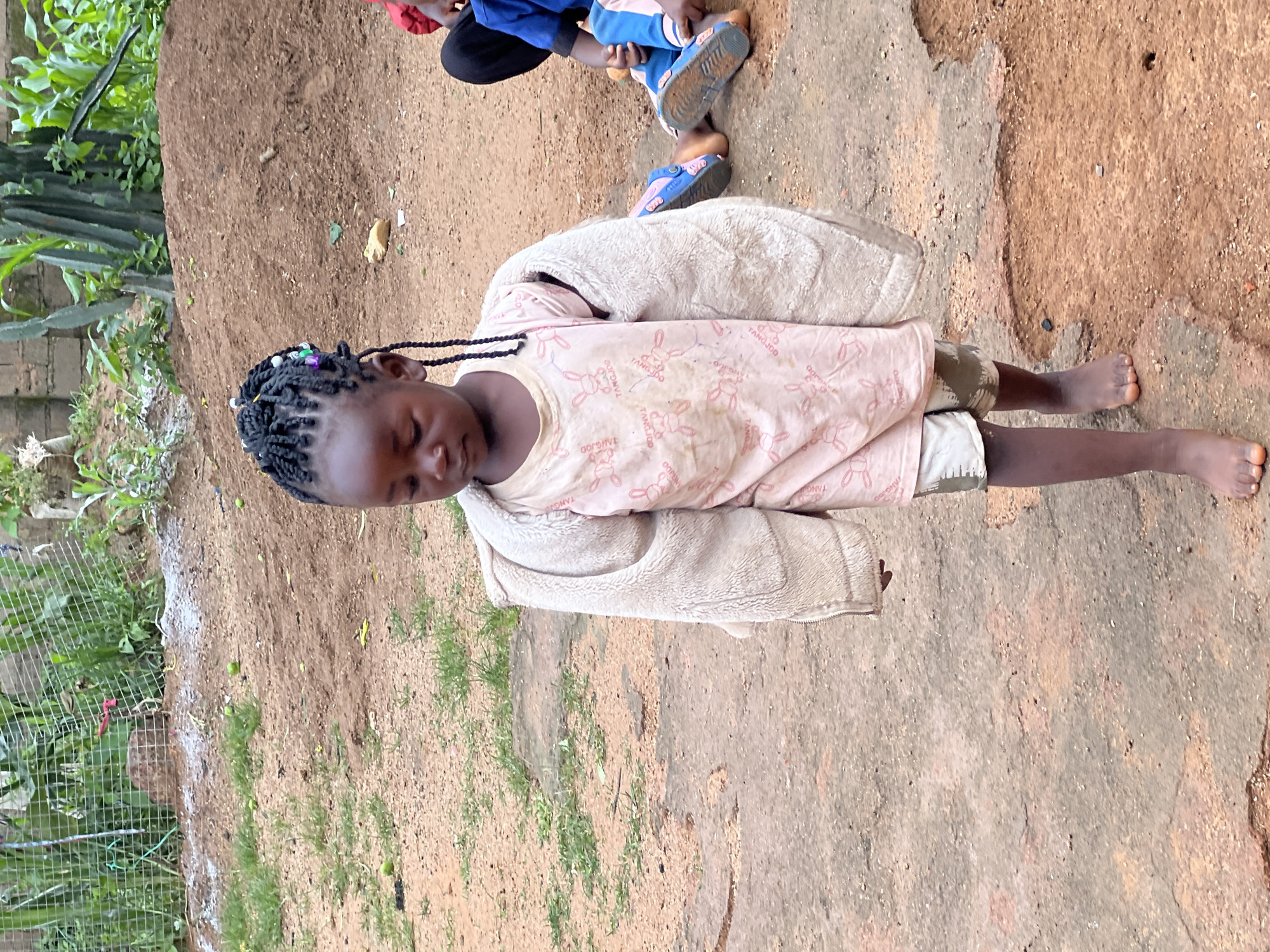 A young Black girl with braided hair adorned with colorful beads walks on a dirt path. She is wearing a light pink t-shirt with a repeating bunny pattern and beige shorts. An oversized, light-colored fuzzy jacket is draped over her shoulders. The girl's expression is somber as she looks down. In the background, two other individuals are seated on the ground, one wearing a blue shirt and the other partially visible in a red garment. They are all in an outdoor setting with dry earth, scattered green vegetation, and a fence in the distance. The environment appears to be a rural or semi-urban area, possibly on the outskirts of Jos, Nigeria, characterized by undeveloped ground and rudimentary structures.