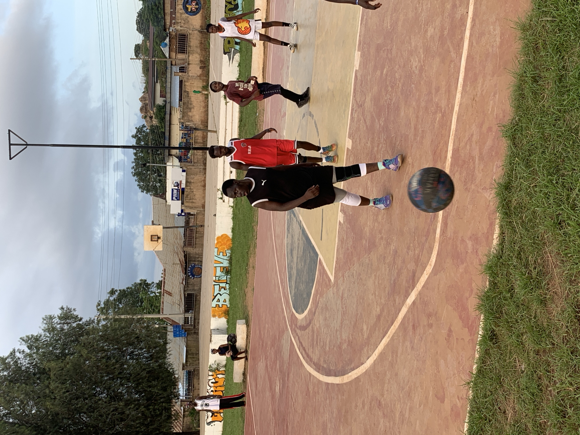 This media file shows a group of young men on a basketball court in Gura Top, Nigeria. The scene is outdoors, with a cloudy sky suggesting either early morning or late afternoon. The basketball court itself is a reddish-brown color with white lines marking the boundaries. To the right of the court, there is a strip of green grass. In the background, there are buildings, some appearing to be shops or residential structures, with corrugated metal roofs. There is also a tall light pole and trees.
Several individuals are visible on the court. One person in the foreground is wearing a black t-shirt and shorts, with white socks and colorful sneakers, and appears to be dribbling a basketball. Another person is wearing a red basketball jersey and shorts. Others are seen in casual clothing, with one person in a white tank top with a flame graphic and white shorts.
Graffiti is present on a wall in the background, with the word "BELIEVE" clearly visible in large, stylized letters, accented with orange. This graffiti, along with the style of the buildings and the presence of a basketball court, suggests a community setting.
The people appear to be engaged in a basketball game or practice. Their stances and the presence of the ball indicate an active sporting event. The lighting suggests it is not midday, and the cloudy sky creates a diffused light. No specific emotions are clearly discernible from the individuals' expressions, but the overall atmosphere seems to be one of casual activity and sports engagement.