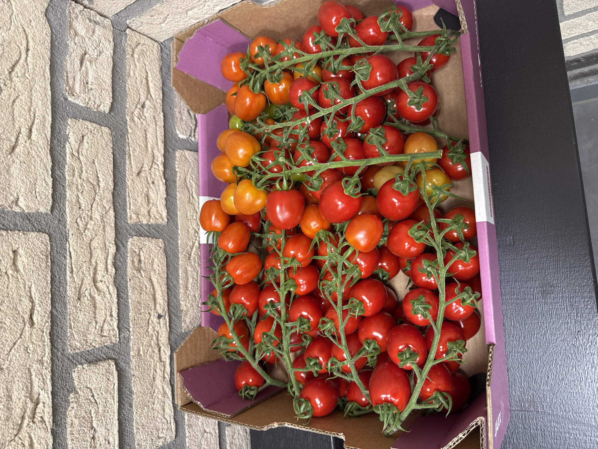 A close-up, overhead shot shows a cardboard box filled with ripe cherry tomatoes on the vine. The tomatoes vary in color from bright red to a deep orange, and some are a vibrant yellow. They are clustered together on green stems, creating a dense, textured surface within the box. The box itself has purple accents and is positioned on a dark surface, possibly a table. In the background, a textured brick wall, likely made of light-colored, rough-hewn bricks with gray mortar, serves as the backdrop. The scene is static and appears to be a still life arrangement, likely in a domestic or retail setting. The location is Den Hoorn, Netherlands.
