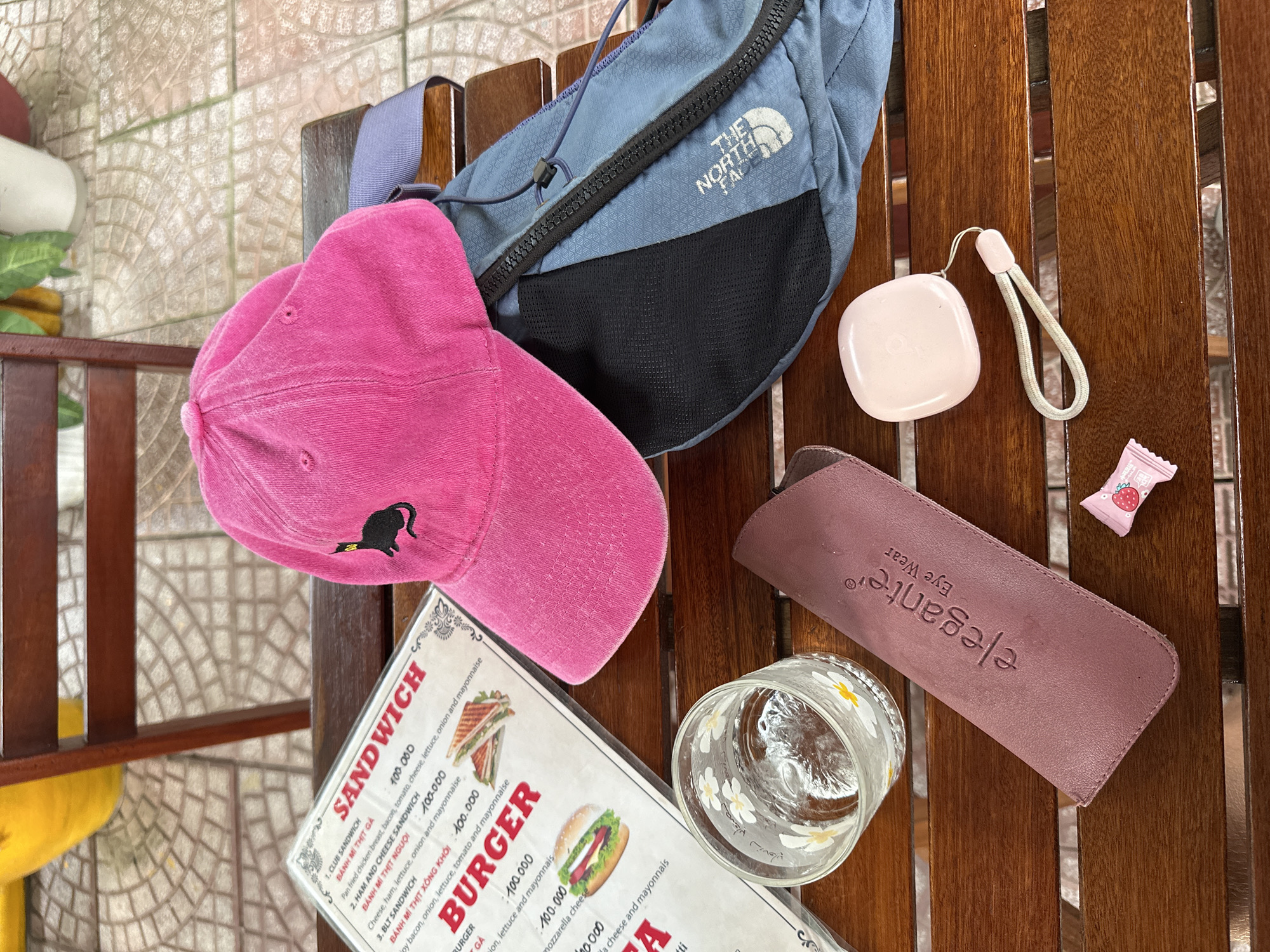 On a wooden slatted table at a cafe in Da Nang, Vietnam, a pink baseball cap with a black cat embroidered on the front lies on top of a blue backpack. A menu is open on the table, listing sandwich and burger options with prices. To the left of the menu is a clear glass with white daisy designs and a bit of water. In front of the glass is a rose-colored glasses case with the word "elegante" and "Eye Wear" printed on it. Next to the case, a small pink candy wrapped in plastic with a strawberry design sits on the table. To the right of the glasses case is a small, square, light pink object with a light gray strap. The background shows a tiled floor and wooden chair legs.