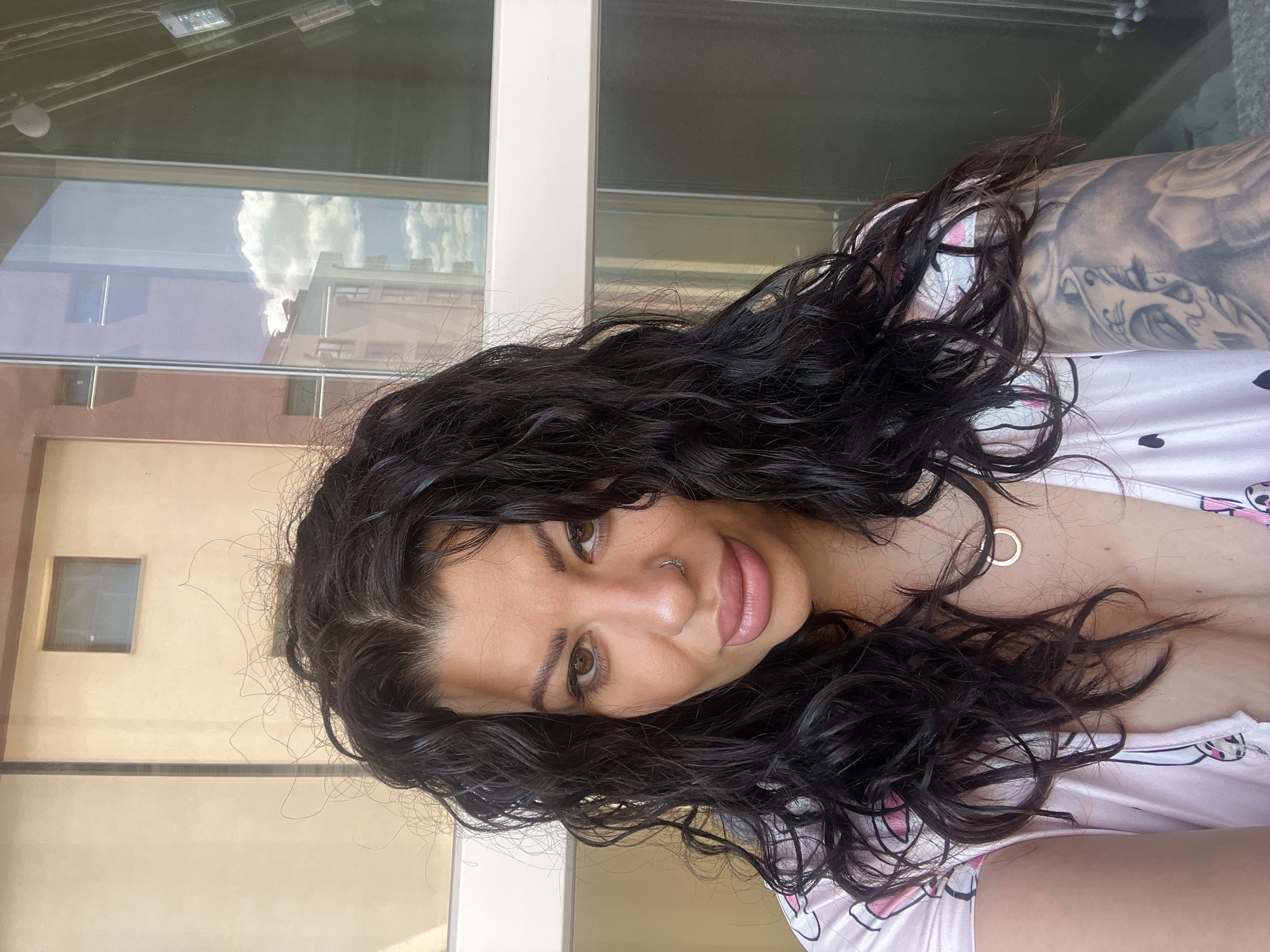 A woman with dark, curly hair and fair skin poses for a selfie in what appears to be an outdoor urban setting. She has a nose ring and is wearing a rose gold necklace. Her left arm shows a large tattoo.
The background is a building with large windows that reflect the sky and nearby buildings. To the left, there is a window that faces the viewer.
Given the urban setting and architecture, the location could potentially be identified further by analyzing architectural styles or visual landmarks unique to Plovdiv, Bulgaria.