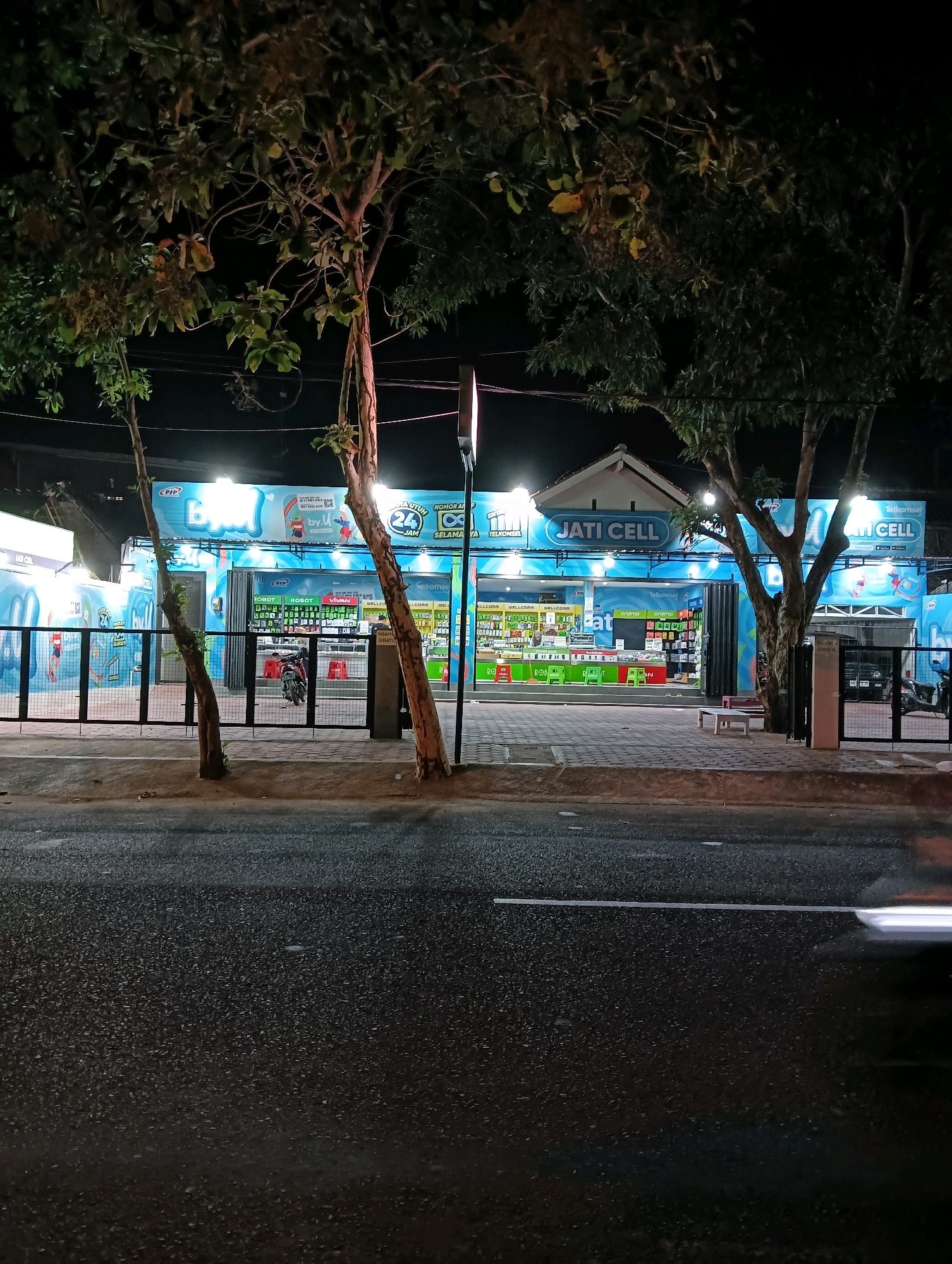 The image depicts a nighttime exterior view of a mobile phone store identified as "Jati Cell" in Purworejo, Indonesia. The store is illuminated by bright overhead lights, showcasing a blue and white facade with prominent signage. Various mobile phone brands and accessories are displayed on shelves visible through the storefront. A black metal gate secures the entrance area, which has a tiled floor. Two young trees with bare branches stand in the foreground, partially obscuring the store. The street in front of the store is paved and appears wet, reflecting the lights from the store and streetlights. A motorcycle is parked inside the gated area, and a small, empty white bench is situated near the entrance. The overall impression is of a commercial establishment operating during evening hours in a town or city setting.