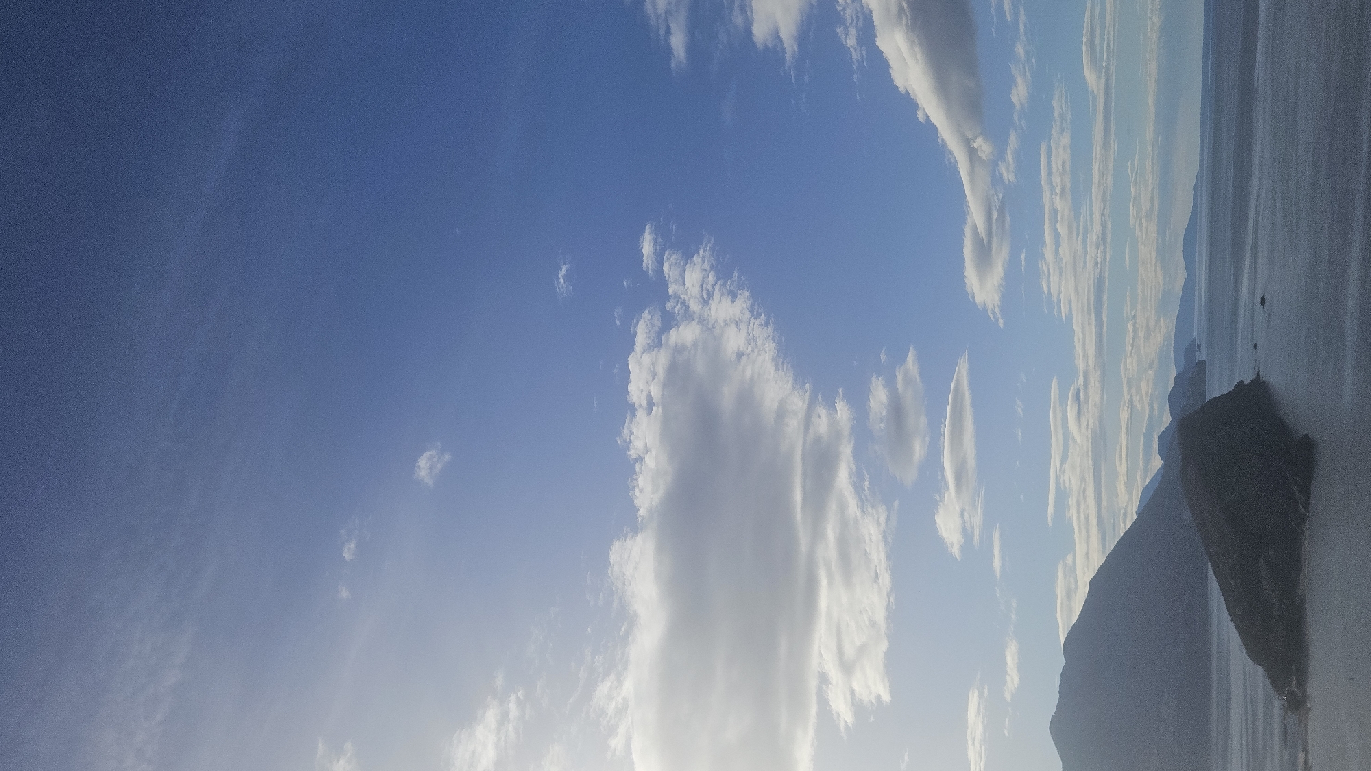 The image shows a landscape with a body of water and mountains under a partly cloudy sky. The sky is blue with scattered white clouds of varying sizes and shapes. In the distance, the body of water extends to the horizon with a series of mountains visible on the right side of the image. Closer to the foreground, a rocky landform is prominent. This location is likely in Budva, Montenegro, given its coastal and mountainous environment. There are no people visible in the image. The focus is on the natural scenery, with the sea, sky, clouds, and mountains as the main subjects.