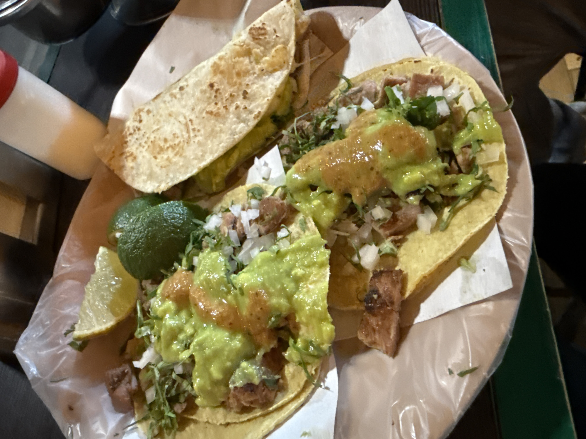 The scene depicts a meal served in San Bernardino Tlaxcalancingo, Mexico. On a light-colored plate, lined with clear plastic and white paper, are three tacos. Two prominent tacos, positioned toward the center and lower right, feature yellow corn tortillas laden with chopped, browned meat, a substantial amount of vibrant green avocado-based sauce, a contrasting drizzle of reddish-orange salsa, finely diced white onions, and green cilantro. A third taco, partially visible in the upper right, utilizes a light brown, griddled tortilla. A whole green lime and a single yellow lime wedge are placed on the plate among the tacos. In the background, a white squeeze bottle with a red cap and a dark metallic container are visible on a dark table surface, which exhibits a green edge in the lower left portion of the image. No individuals are visible.