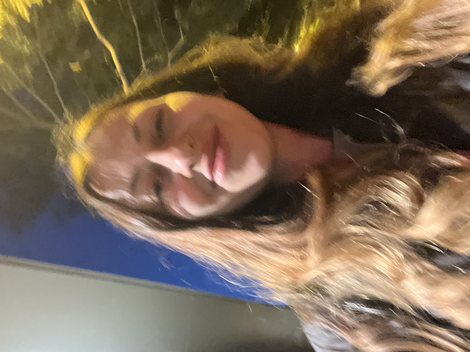 A selfie-style, slightly blurry photo depicts a woman with long, wavy hair that transitions from dark roots to a lighter, golden-brown hue. She is positioned at an angle, and the focus is on her face, which appears softly lit. The background is a blend of blue and light grey suggesting the sky and a building, while yellow light suggests street lighting and/or signage overhead. While not explicitly apparent from this photo, the specification is that the location is Barcelona, Spain.