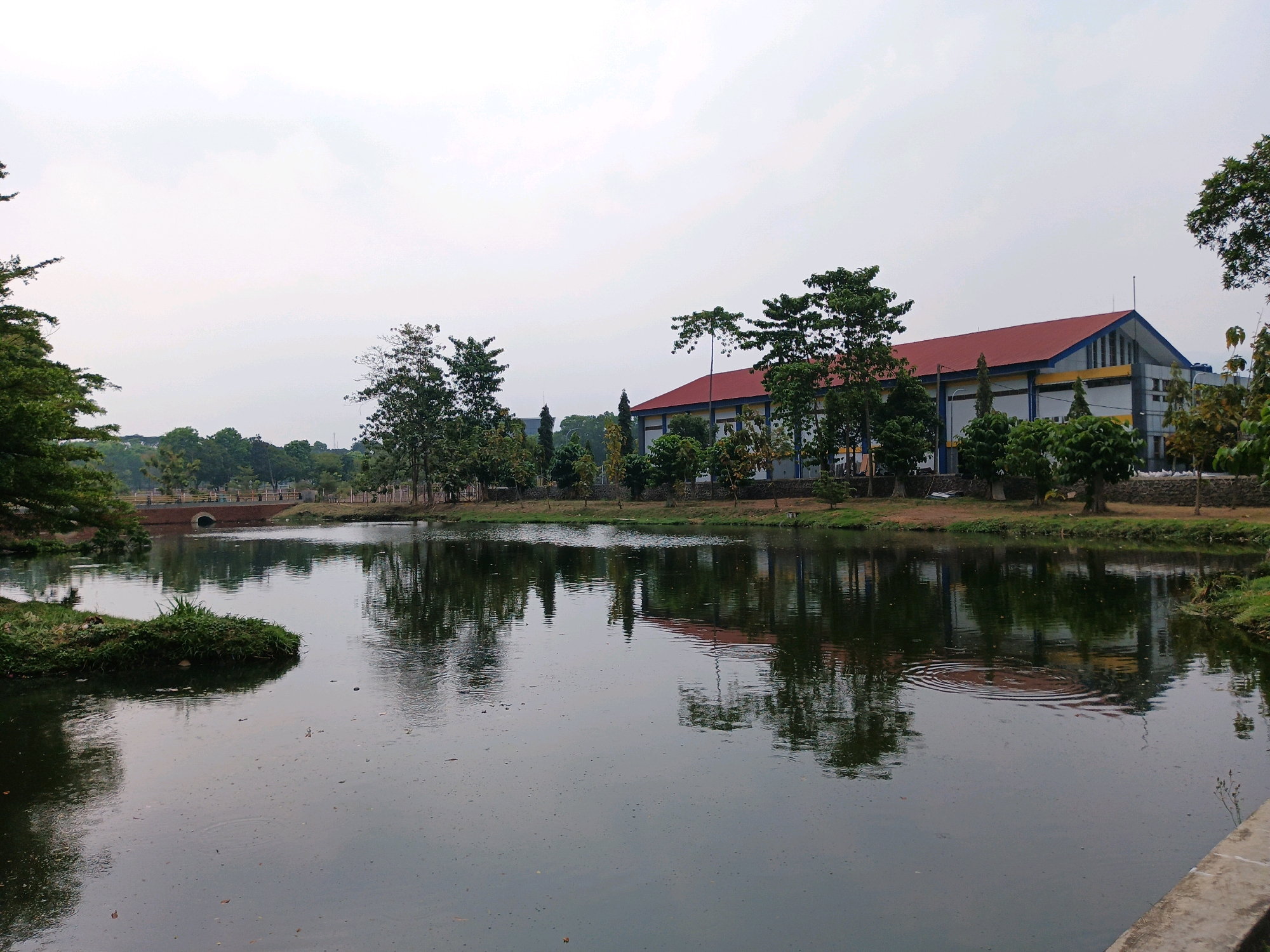 This image shows a tranquil scene with a pond in the foreground, reflecting trees and a building. The building appears to be a sports facility or gymnasium, characterized by its red roof and light-colored walls with blue and yellow accents. The surrounding environment includes lush green trees and vegetation, a pathway, and a small arched bridge in the distance. The sky is overcast, contributing to the calm and muted atmosphere of the scene. The overall impression is that of a well-maintained public space, possibly a park or a campus in Bandung City, Indonesia, offering a serene escape within an urban setting.