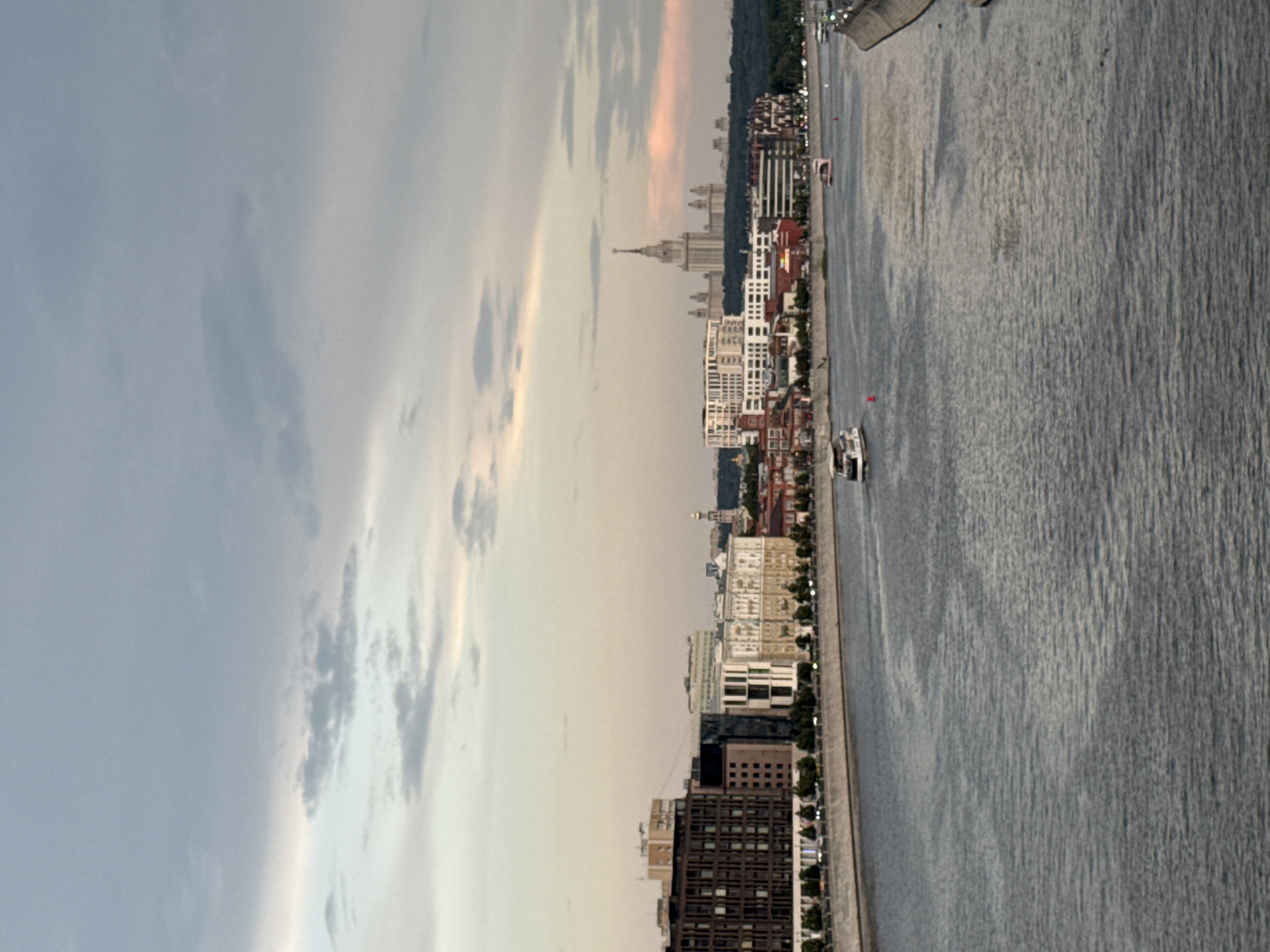 The image depicts a panoramic view of the Moscow River and its embankment in Moscow, Russia. The sky is overcast with soft, diffused light, suggesting it is either early morning or late evening. Several boats are visible on the water, with one in the foreground and others further in the distance.
The embankment is lined with trees and modern buildings of varying architectural styles. On the left, a dark, multi-story building with a patterned facade dominates the scene. To its right, a cluster of buildings includes structures with white and beige facades, some featuring prominent window arrangements. Further down the embankment, more residential or office buildings can be seen.
In the background, a large, ornate Stalinist skyscraper, the main building of Moscow State University, stands tall, its spire piercing the sky. The presence of this iconic landmark, along with the Moscow River and the characteristic architecture, firmly places the scene in Moscow. The overall impression is of an urban landscape with significant architectural features and active waterways.