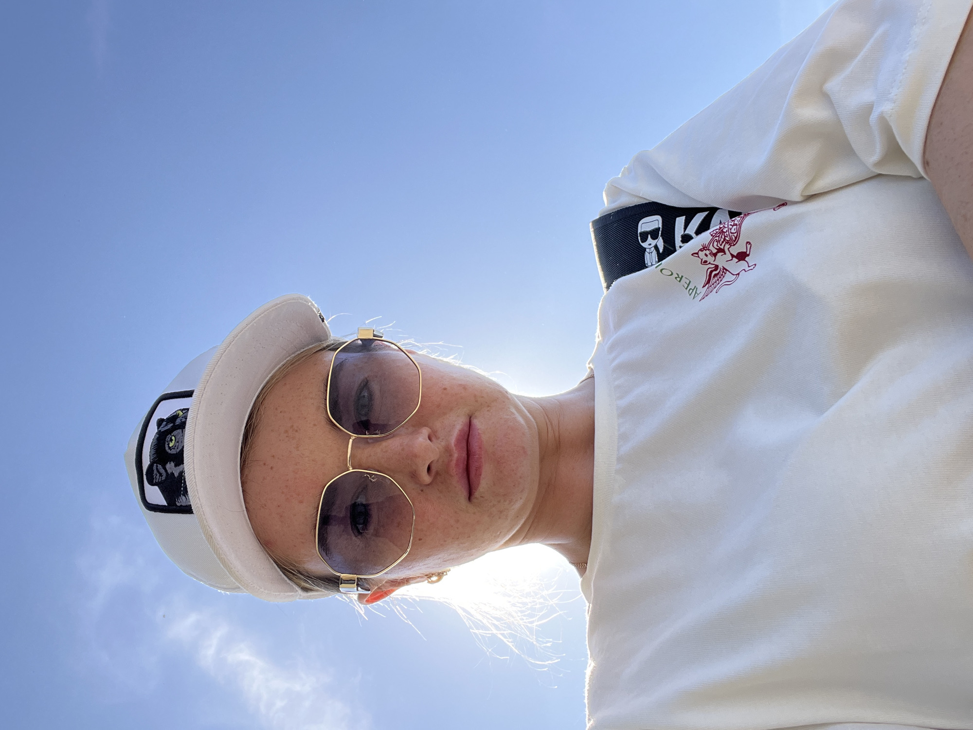 A woman is photographed from a low angle against a bright blue sky. She is wearing a white cap with a black patch depicting a gorilla, and sunglasses with gold frames. Her face is visible, showing freckles. She also wears a white t-shirt with a red design on the chest. A black strap with a cartoon figure can be seen over her shoulder. The location cannot be determined to be Chaikovsky, Russia from the image.