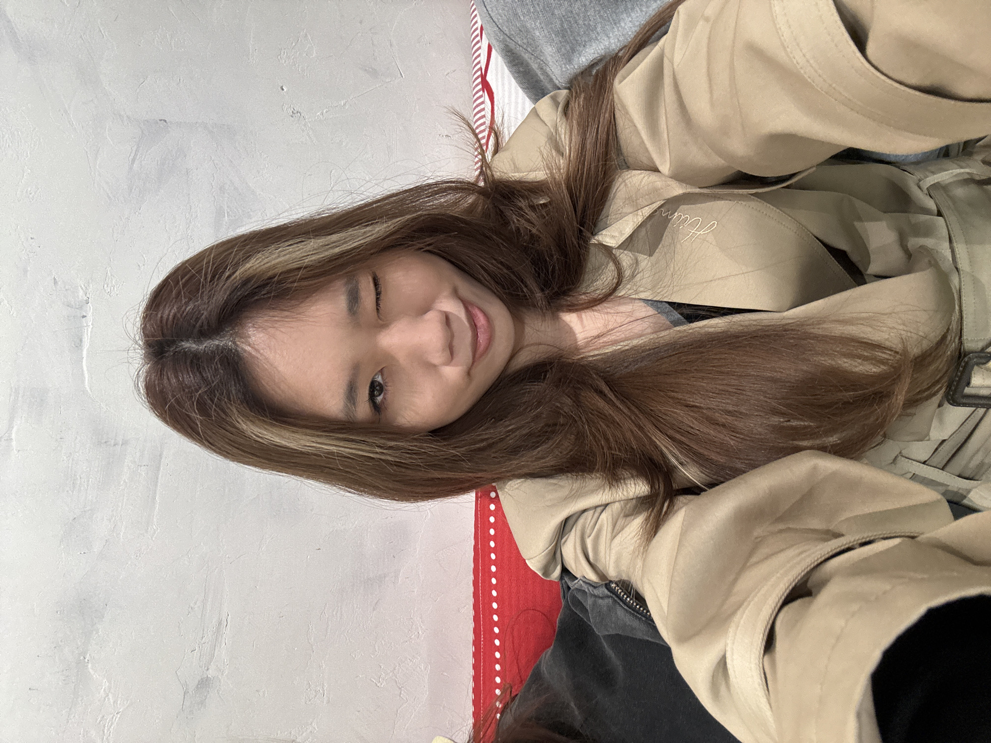 A young woman with long brown hair and a blonde streak is winking at the camera. She is wearing a tan jacket and sitting on a surface with red and white stripes. The background is a white wall. The location of the image cannot be determined from the visual information provided. The mention of Jianlan Road District, China, is not relevant to the visible content of the image.