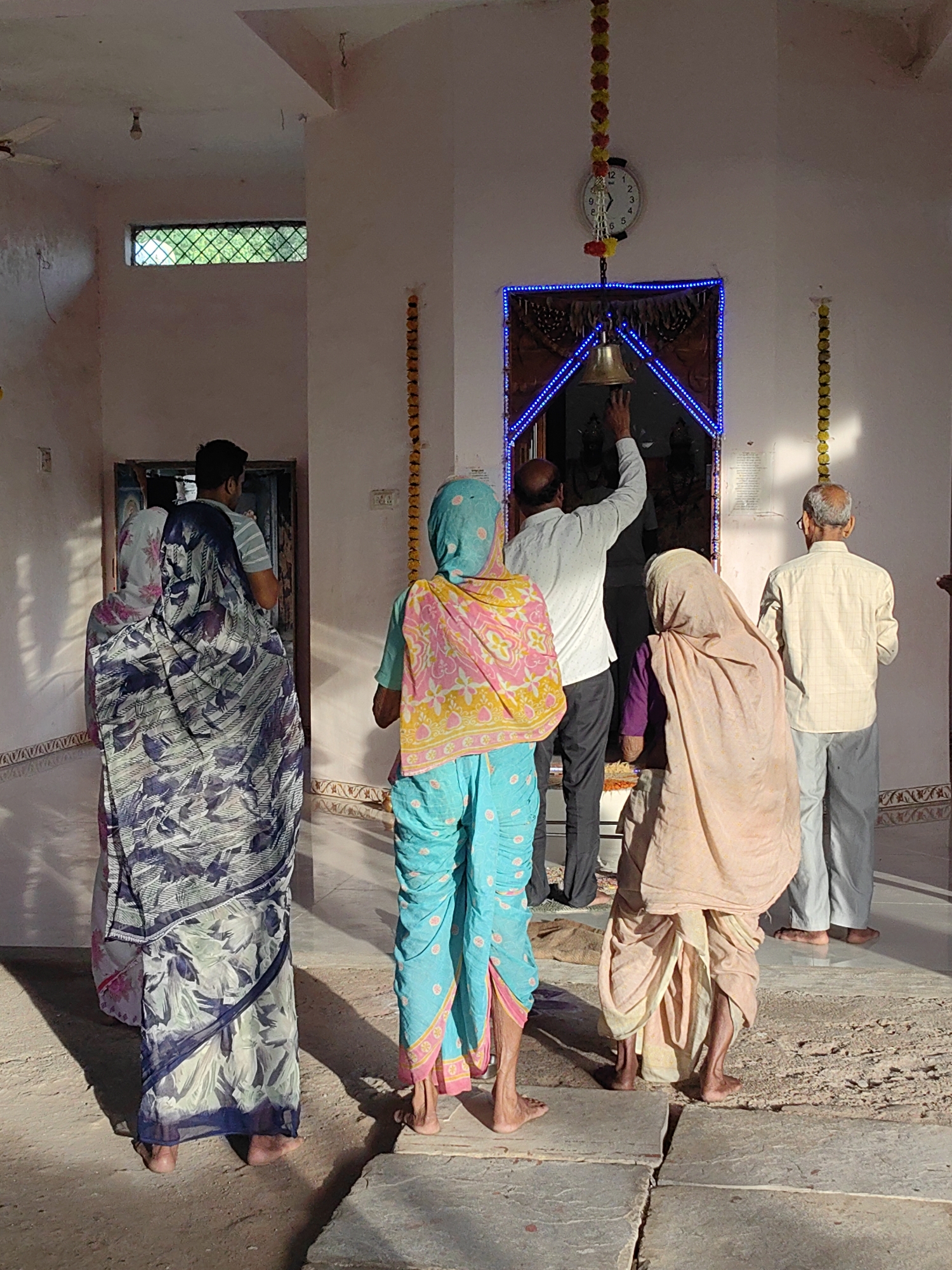 In Mhaiswadi, India, a group of people are gathered inside a building, likely a temple or shrine. The lighting suggests it is daytime, with sunlight casting distinct shadows on the floor. Several individuals are facing towards an entrance adorned with blue lights and a hanging bell, which one person is reaching out to touch. The architecture features a light-colored, possibly pink, interior with a clock on the wall and decorative garlands. The people are dressed in traditional Indian attire, including saris and kurtas. Some are barefoot, while others wear footwear. The scene conveys a sense of religious observance or a communal gathering.