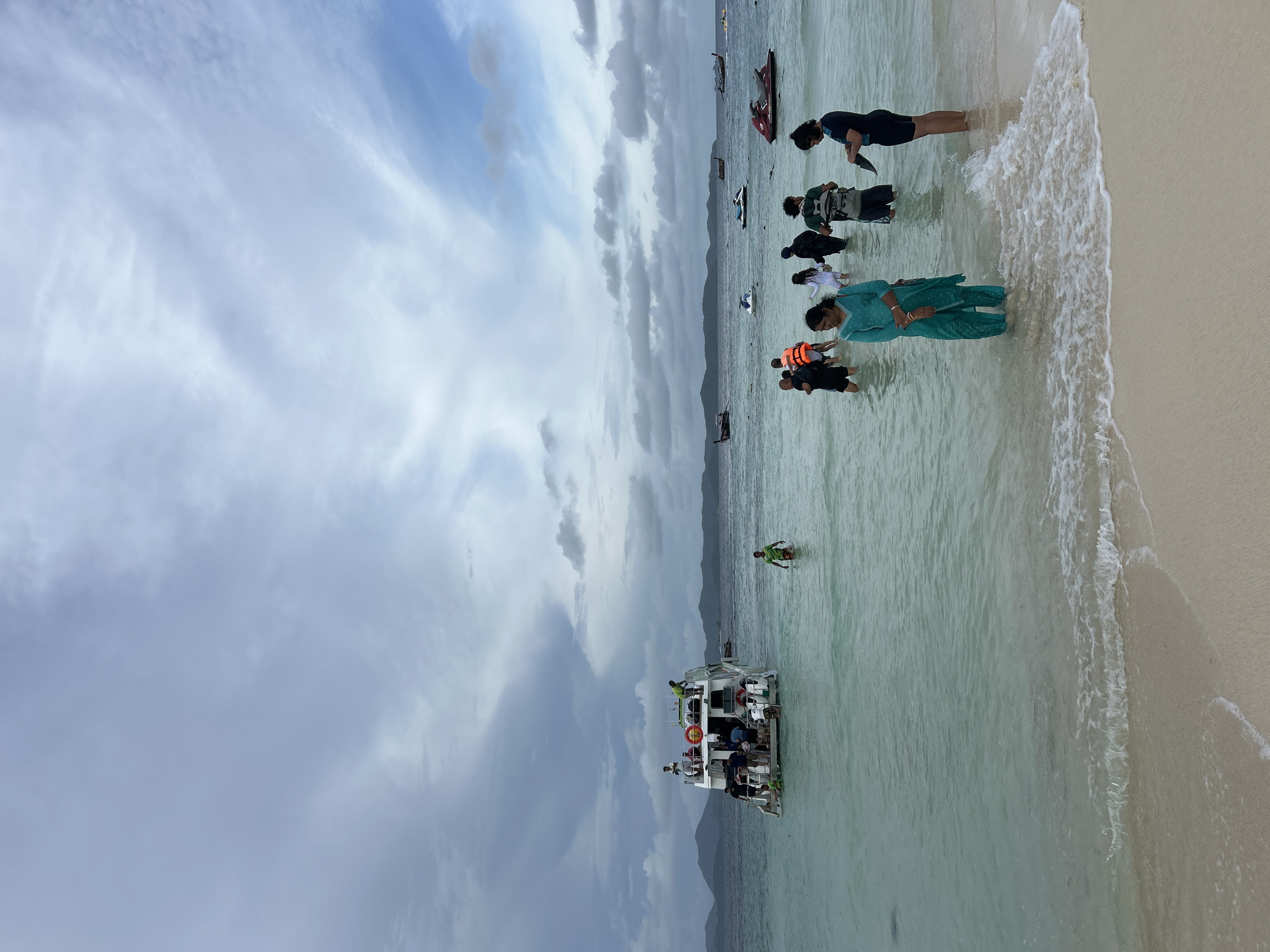 In Tambon Phru Nai, Thailand, on a cloudy day, several people are wading in the shallow, clear turquoise waters of the sea near a sandy beach. A large white tour boat with multiple levels is anchored in the water, carrying passengers. Other smaller boats, including jet skis, are visible further out in the sea. The people on the shore and in the water are casually dressed, with some wearing life vests. The water is calm, with gentle waves lapping at the shore. The background shows a hazy, mountainous coastline.