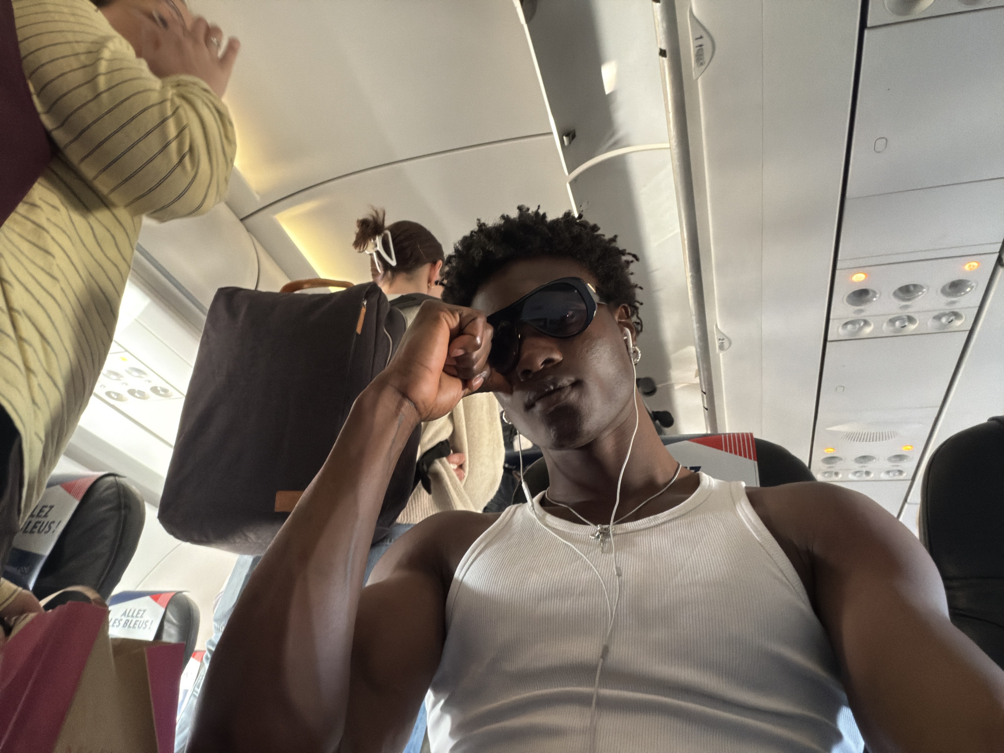 A young Black man takes a selfie inside an airplane cabin, occupying the central foreground of the image. He wears dark sunglasses, a white ribbed tank top, and wired earbuds, with a silver chain around his neck. His right hand is resting near his face, showing muscular definition in his arm.
The setting is an airplane interior, characterized by overhead compartments, adjustable air vents, reading lights, and seatbelt signs, some of which are illuminated amber. Other passengers are visible in the background; to the left, a person in a striped long-sleeved shirt is partially seen, and further back, another person with dark hair tied in a bun is largely obscured by a large dark tote bag.
The cabin appears to be active, suggesting boarding or disembarking. Paper shopping bags, one pink and one light brown, are visible near the front-left seat.
Notable details include the text "ALLEZ LES BLEUS!" prominently displayed on the headrest covers of multiple seats, which is a common French sports chant. An overhead panel also shows partially visible text that appears to be "1 rang" (French for "1 row"). These linguistic cues strongly suggest a French airline or a flight related to France. The specific location of Crawley, United Kingdom, is not visually discernible from the image itself. The time of day is not clear, but the cabin is well-lit by internal lighting and some ambient light. The main subject's expression behind the sunglasses appears cool or neutral.