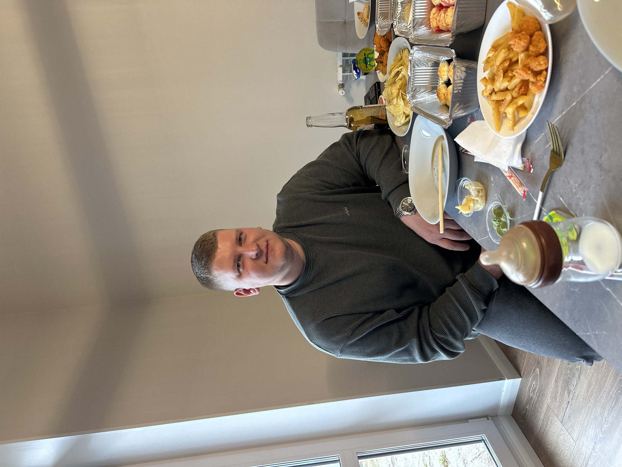 The image shows a man sitting at a table laden with food. The table has dishes of chips, fried food, and dipping sauces. A beer bottle and baby bottle are also present. The man is wearing a dark green sweater and a watch. He is looking at the camera. The background shows a wall and a window with a view of trees. The location is a room in what appears to be a house, not identifiable as being in Chaikovsky, Russia without additional information.