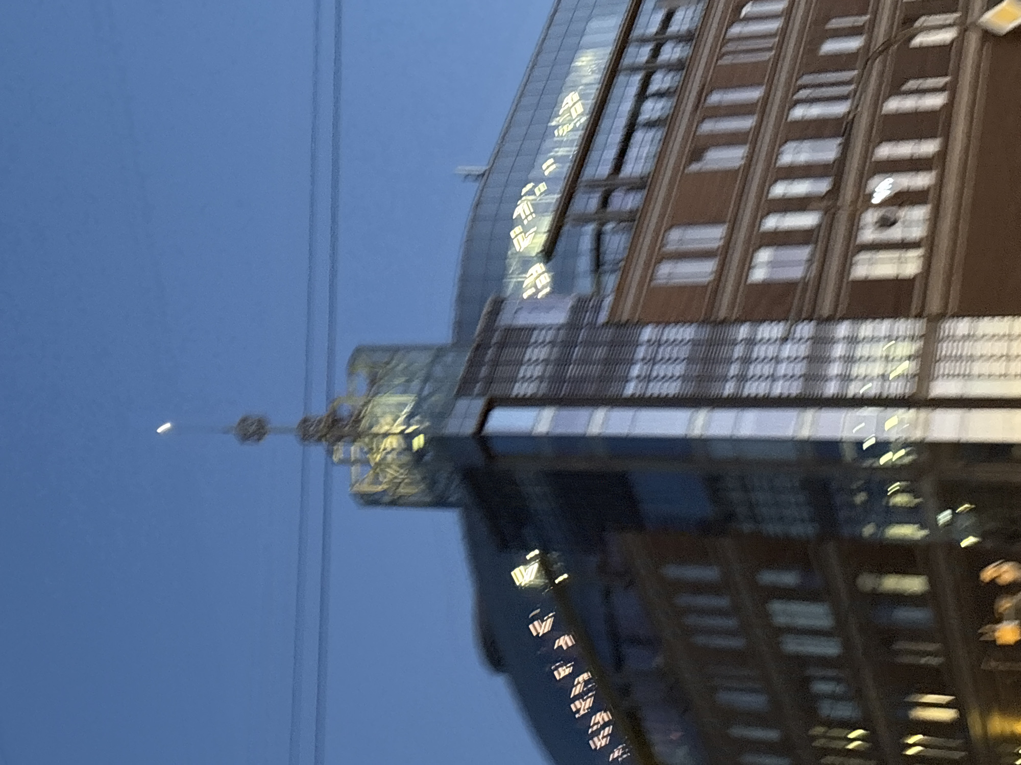 The image shows a modern building with a glass facade reflecting the blue twilight sky and lights from within. The building has a brick lower section and a curved glass roof. At the very top of the building, a decorative spire is visible. The image is taken in the city of Saint Petersburg, Russia. A thin crescent moon is visible in the sky, positioned above the building's spire. Power lines are stretched across the upper portion of the image. The overall scene suggests an urban environment at dusk or early night.