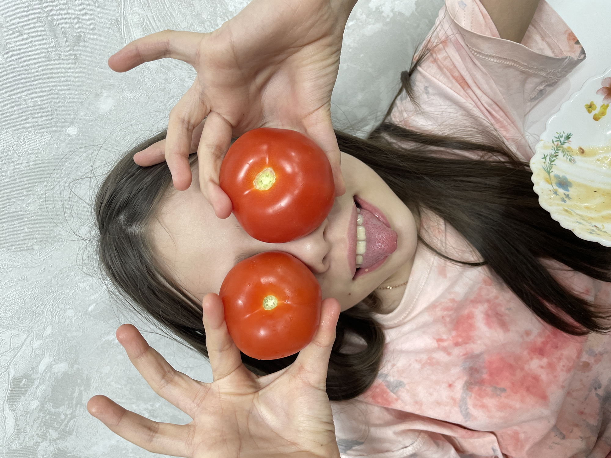 A woman lies on her back, holding two red tomatoes over her eyes. She has dark hair and is wearing a pink tie-dye shirt. Her mouth is open, and her tongue is sticking out. One hand is above her head, and the other is below. To the right of her head, there is a plate with remnants of food on it. The background is a light-colored wall. There is no evidence to suggest this photograph was taken in Izhevsk, Russia.