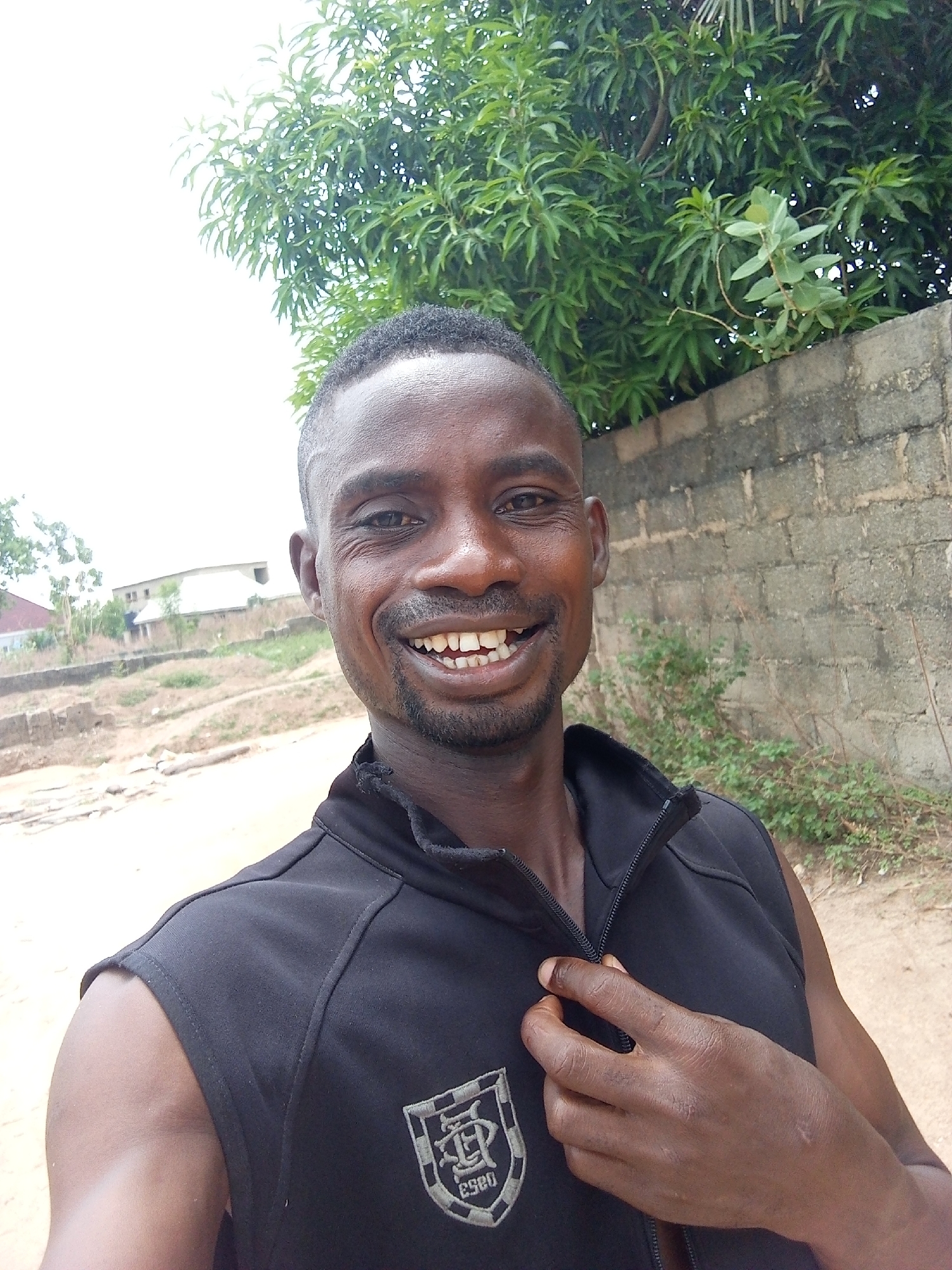 The image is an outdoor, close-up portrait of a dark-skinned adult man, likely taken as a selfie or by someone close to him. The man has short hair and a beard, and he is captured with a wide, open-mouthed smile, showing his teeth and conveying a happy and jovial expression. He is wearing a black sleeveless vest or jacket with a central zipper and a distinctive grey and white shield-shaped logo on the left chest. The logo features stylized letters, possibly "DP," and the number "2020" at the bottom. His right hand is visible, lightly touching the zipper of his vest.
The background suggests a daylight setting, likely under a bright sky. To the man's right, a grey concrete block wall is visible, partially covered by sparse green shrubbery. Above the wall and extending into the upper right of the frame is a large tree with abundant green foliage. To his left, the background reveals a dusty or unpaved dirt path, leading towards some simple, light-colored buildings in the distance under a bright, possibly overcast, sky. The scene depicts a semi-urban or rural environment consistent with a location like Adonkolo, Nigeria.