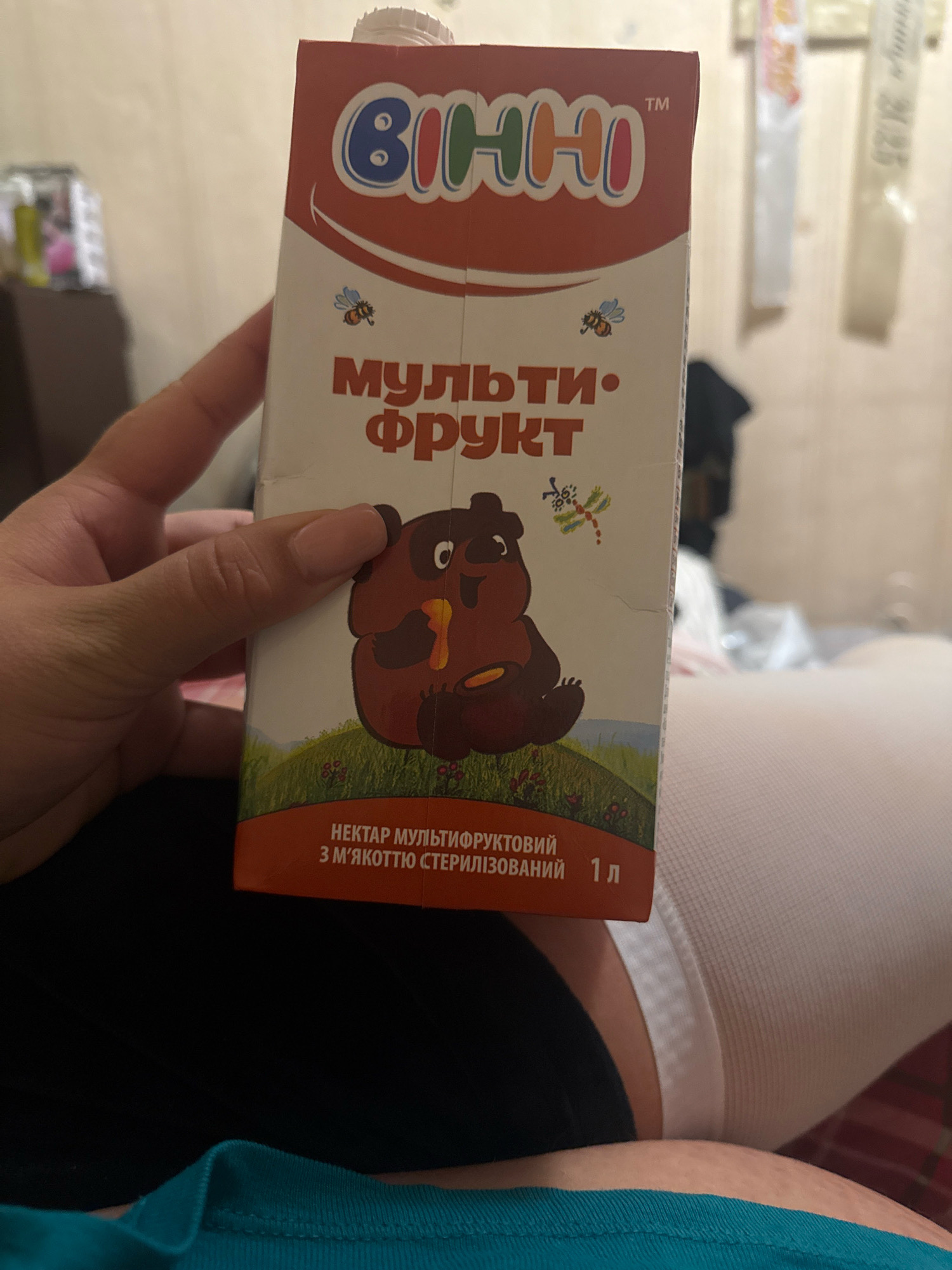 A person's hand holds a carton of "Bi-Oh-No" brand multi-fruit nectar in the city of Kuna, Ukraine. The carton features a cartoon of a bear holding a pot of honey and a dragonfly. The text on the carton is in Ukrainian and indicates that it is a sterilized nectar with pulp. The background is blurred and appears to be an indoor setting, possibly a bedroom, with light-colored walls and furniture. The person is wearing a dark-colored garment and a white compression sleeve on their leg.