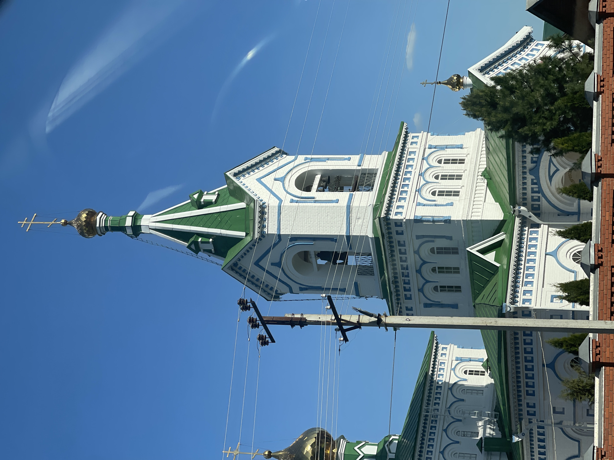 The image depicts a church in Перевозное, Russia, with white walls, blue trim, and a green roof. A golden dome topped with a cross rises above the structure. An adjacent brick building is partially visible. A utility pole with power lines bisects the view, contrasting with the clear blue sky in the background.