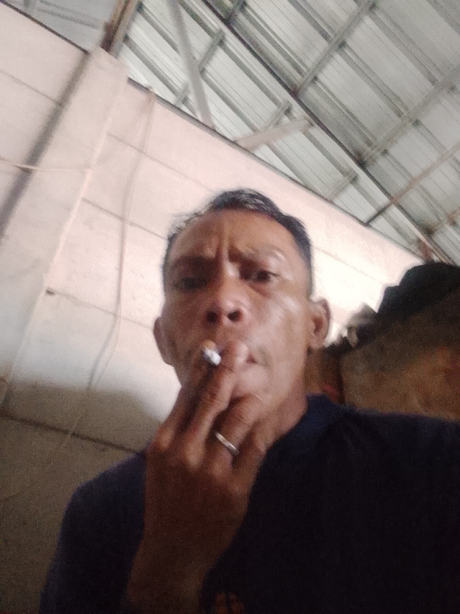 A man is holding a lit cigarette between his fingers, near his mouth, likely inhaling or about to inhale. He is wearing a dark-colored shirt and a wedding band on his finger. The setting appears to be an interior with corrugated metal roofing and exposed structural elements. The wall is made of concrete blocks. The image was taken in Bukit Ketok, Indonesia.