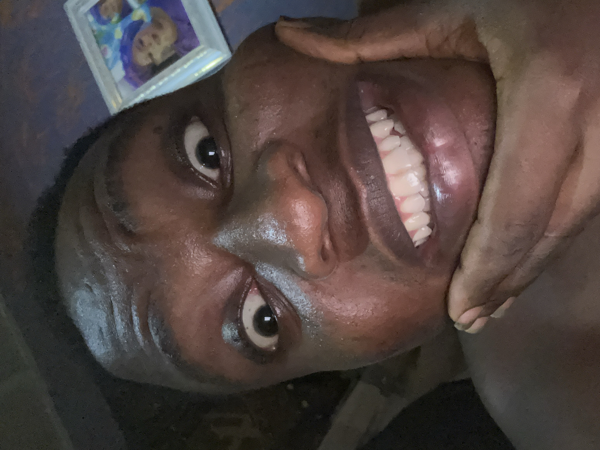 The image depicts a close-up, low-angle view of a dark-skinned person's face. The person is smiling widely, exposing their teeth. A hand is holding the person's chin. A framed photo with faces is visible in the upper left corner. Due to the lack of specific location details within the image itself, it is impossible to confirm its location as Ikorodu, Nigeria. There is no evidence of a cityscape or any specific landmark. The description is based solely on the visual information within the provided image.