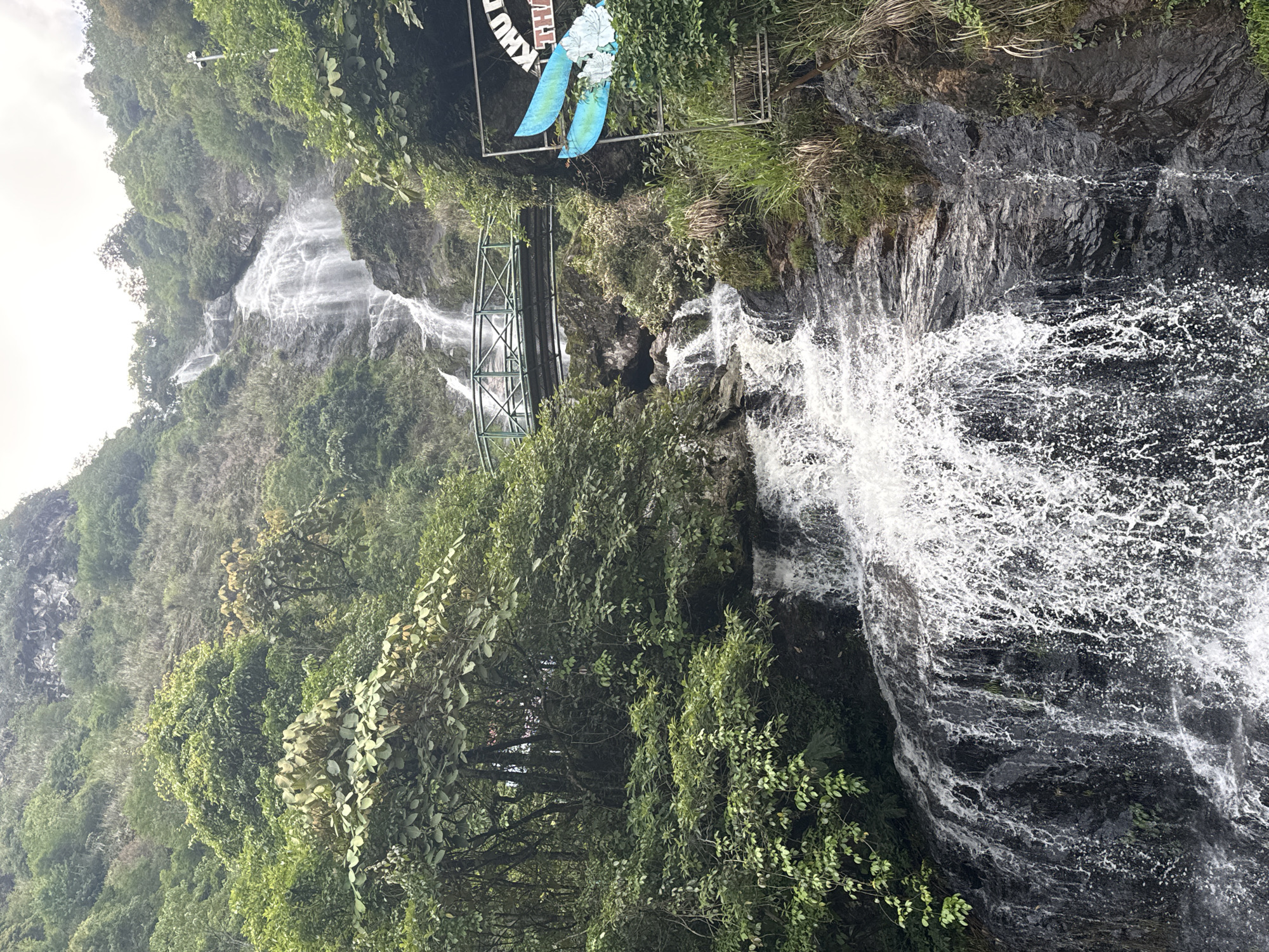 The image captures a waterfall scene, likely in the vicinity of Sa Pa Ward, Vietnam, given the lush greenery and mountain setting. The waterfall itself has two visible sections. A distant upper section cascades down a rocky mountain slope covered in dense vegetation. A closer, more immediate section is characterized by a powerful rush of water cascading over dark rocks. The spray from this section creates a white mist.
A metal bridge structure, partially obscured by foliage, spans a part of the waterfall. Above the bridge is a sign, which is illegible but appears to be a park or tourist area marker. Dense trees and green foliage cover much of the surrounding area, creating a dense, humid environment. The image is taken from an elevated position. The sky is overcast.