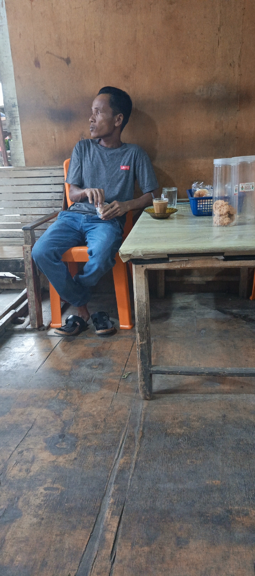 In a dimly lit, rustic establishment in Garot, Indonesia, a man sits alone at a wooden table. He is wearing a grey t-shirt with a red Levi's logo and blue jeans. His feet are clad in black sandals. The man is holding a small, wrapped item in his hands, appearing to be in the process of unwrapping it or taking a bite.
Before him on the table, a small glass of a brown beverage, possibly coffee or tea, sits on a saucer. Next to it is a blue basket containing a single, wrapped pastry. To the right, a large clear plastic container, possibly a dispenser for snacks, holds several large, round, cookie-like items. The table itself is covered with a light-colored, possibly plastic, tablecloth.
The man is seated on an orange plastic chair, its bright color contrasting with the muted tones of the surroundings. The background consists of a wooden wall, possibly unfinished or weathered, with visible grain and some darker smudges. To the left, a slatted wooden structure, possibly a bench or part of the building's construction, is partially visible. The floor is made of worn wooden planks with a distressed, aged appearance. The overall atmosphere suggests a casual, perhaps roadside, eatery or rest stop.