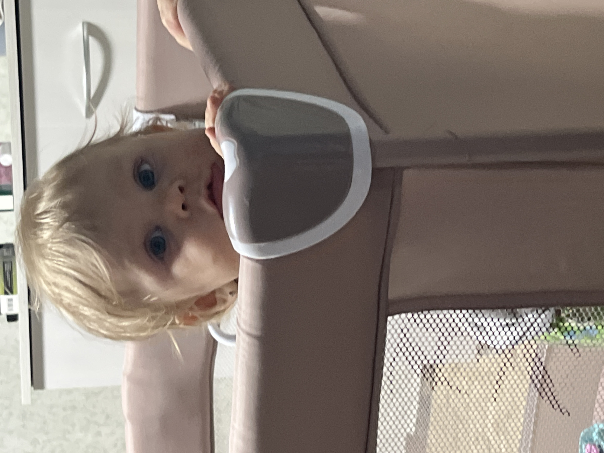 The image displays a blonde-haired, blue-eyed child, appearing to be a toddler, positioned inside a beige or light brown playpen. The child's head and upper torso are visible, with both hands resting on the top padded edge of the playpen. A grey and white curved plastic guard is present on the playpen's top right corner. The child's gaze is directed forward. In the background, a white cabinet or dresser with a silver handle is visible, upon which several small items are present, including a black and a green object. The wall behind the furniture features patterned wallpaper. A mesh panel of the playpen is discernible in the lower-left quadrant of the image. The scene is an indoor environment within a residence in the city of Фоки, Russia.