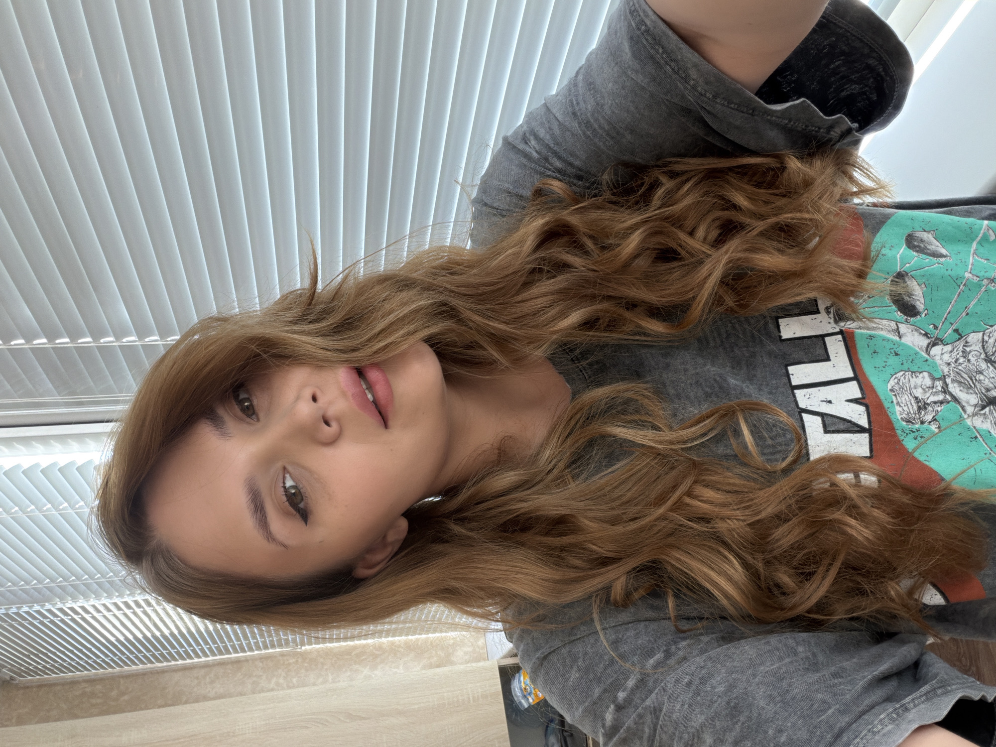 A woman with long, light brown, wavy hair is posing for a photo. She has fair skin, light pink lips, and eyeliner on her eyes. She is wearing a grey hoodie and a t-shirt with a design on it. The background includes a window with white blinds and a light-colored wall and desk. The location is unknown but is said to be in the city of Chaikovsky, Russia.