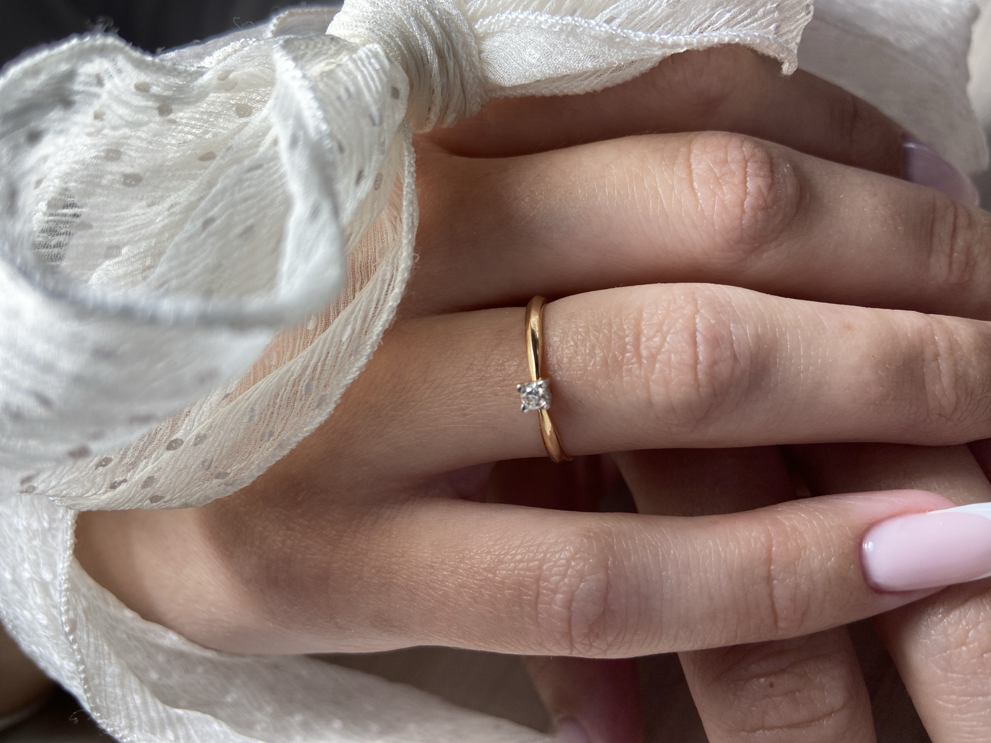 The image depicts a person's hands with a gold ring on the finger. The ring has a small diamond setting. The fingers are intertwined, and a white, textured fabric is draped around the hands. The nails are manicured with a pink base and white tips (French manicure). No specific location in Budva, Montenegro can be determined from this image.