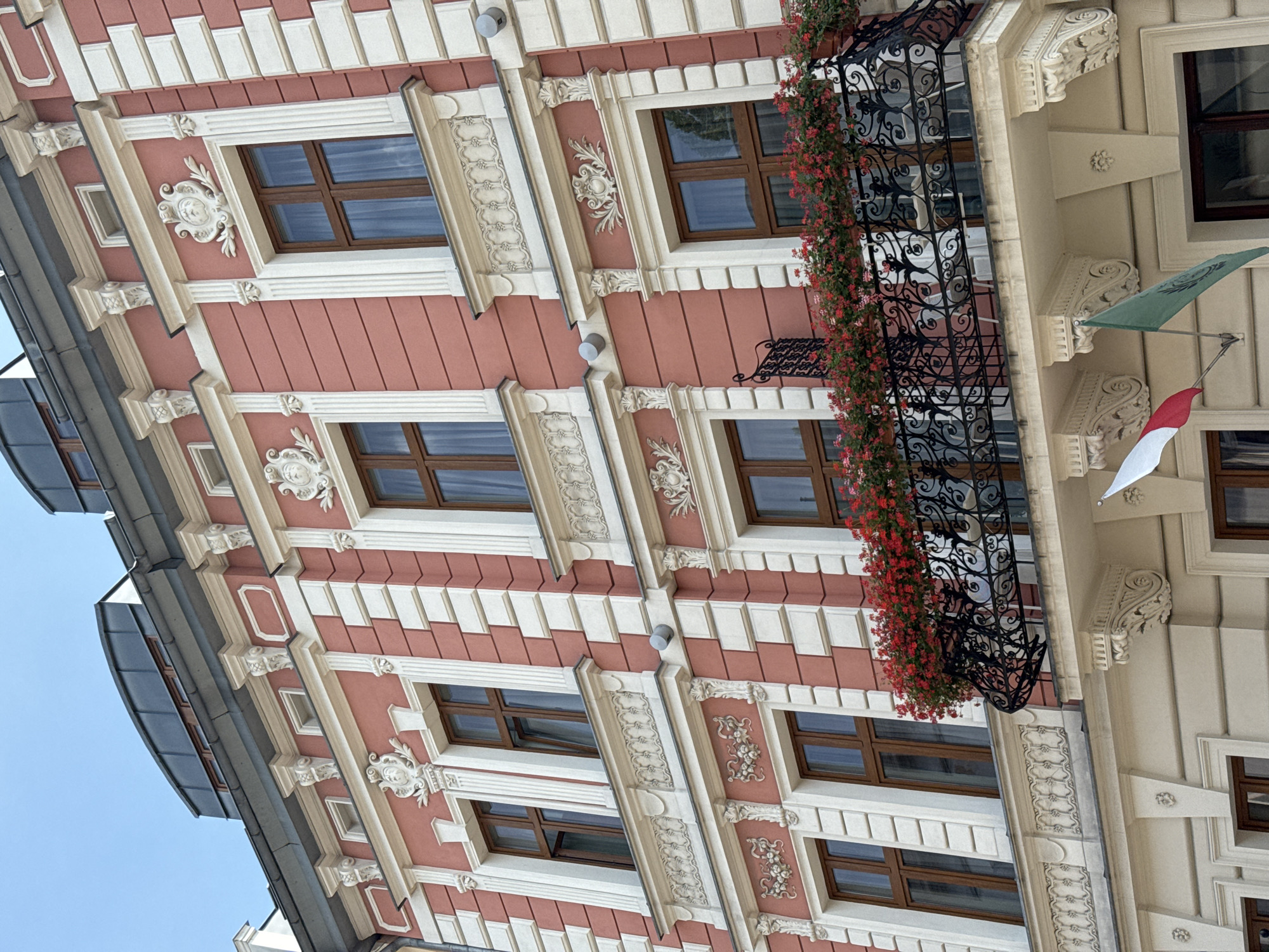 This image shows a close-up, low-angle view of the facade of a historic building in Krakow, Poland. The building has a coral pink and cream color scheme with ornate white plaster detailing around the windows and cornices. The windows are tall and rectangular with dark brown frames. A black wrought-iron balcony, overflowing with red flowers, extends from the second story. Two flags, one red and white and the other green, are visible hanging from the building on the lower level. The sky above is a clear blue. The architecture suggests a style common in the Old Town or Kazimierz district of Krakow.