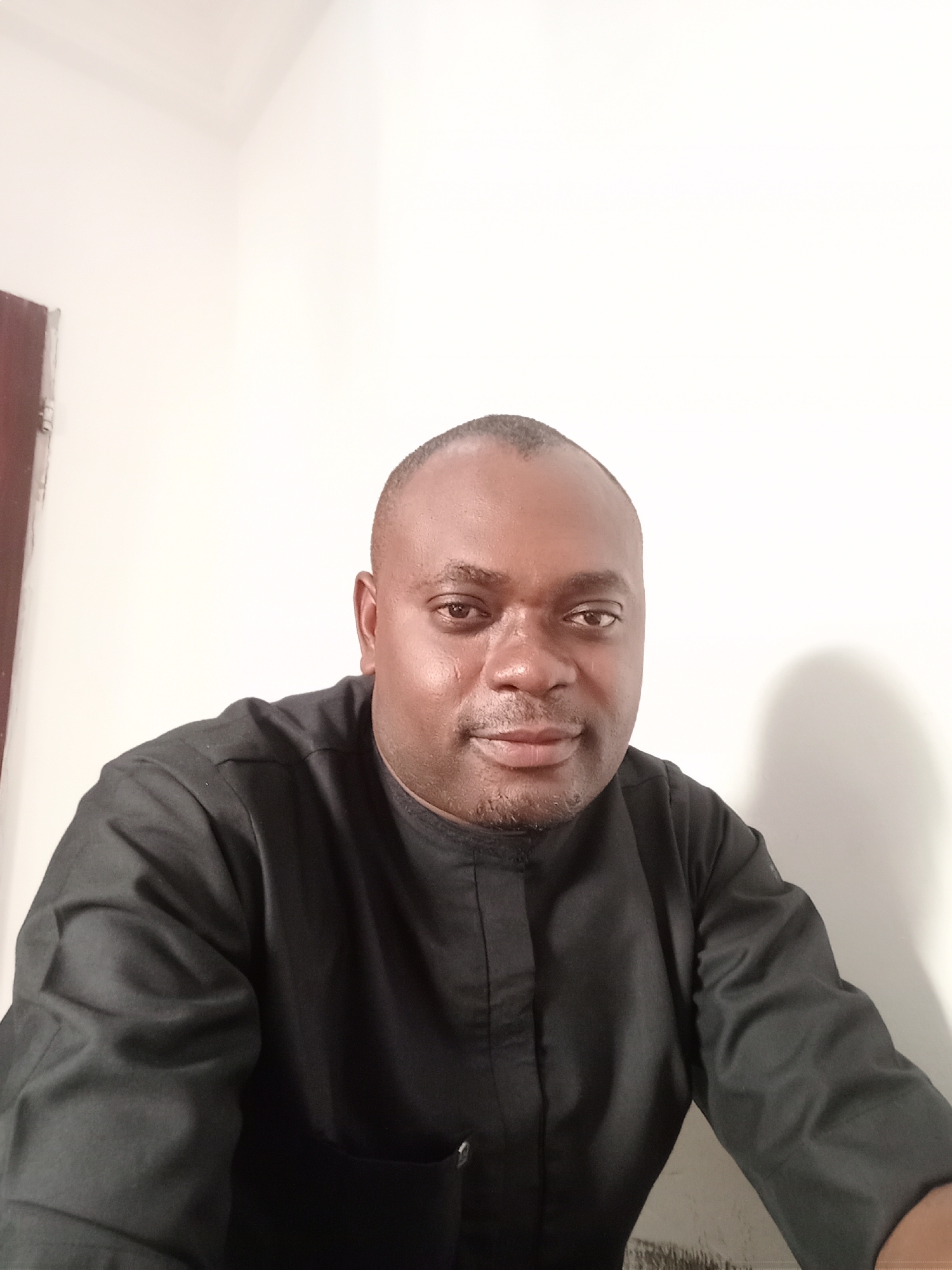 A headshot-style portrait captures a dark-skinned adult male indoors, looking directly at the camera. He appears to be in his 30s or 40s, with a shaved head and short, dark stubble or a neatly trimmed beard. The man wears a long-sleeved black traditional garment, possibly a kaftan or similar tunic, which covers his torso. His expression is calm and neutral, featuring a subtle, closed-mouth smile and engaged eyes, conveying a sense of confidence and relaxation.
The background is a plain, light-colored wall, likely white or off-white. On the far left of the frame, a narrow vertical strip of a dark brown door or doorframe is visible. A soft shadow is cast on the wall behind and to the right of the man, indicating an indoor setting with ambient lighting, likely during the daytime. The minimalist background focuses attention on the subject. The photograph appears to be a posed portrait. No visible text is present. The context provided suggests the location is Gwarinpa, Nigeria.