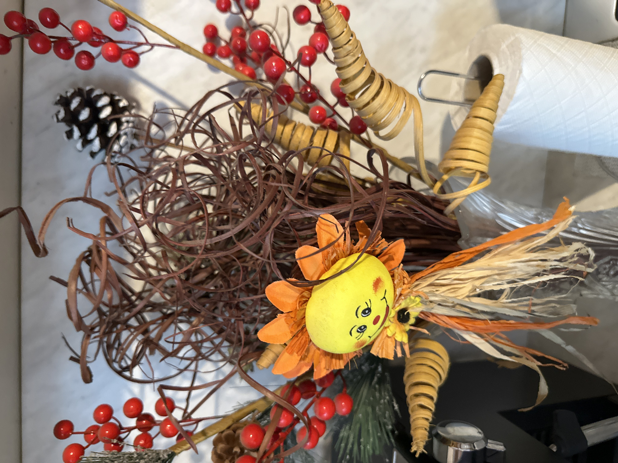 The image shows an overhead view of a decorative arrangement. It consists of a yellow, egg-shaped face with painted features surrounded by orange fabric petals. Brown, twig-like elements are clustered around the face, interspersed with red berries on stems, and spiraled, cone-shaped decorations. A pine cone and white paper towels are partially visible. The arrangement appears to be on a countertop. There is no indication of a location in Montana, Bulgaria, or any specific city. No people, actions, or interactions are depicted.