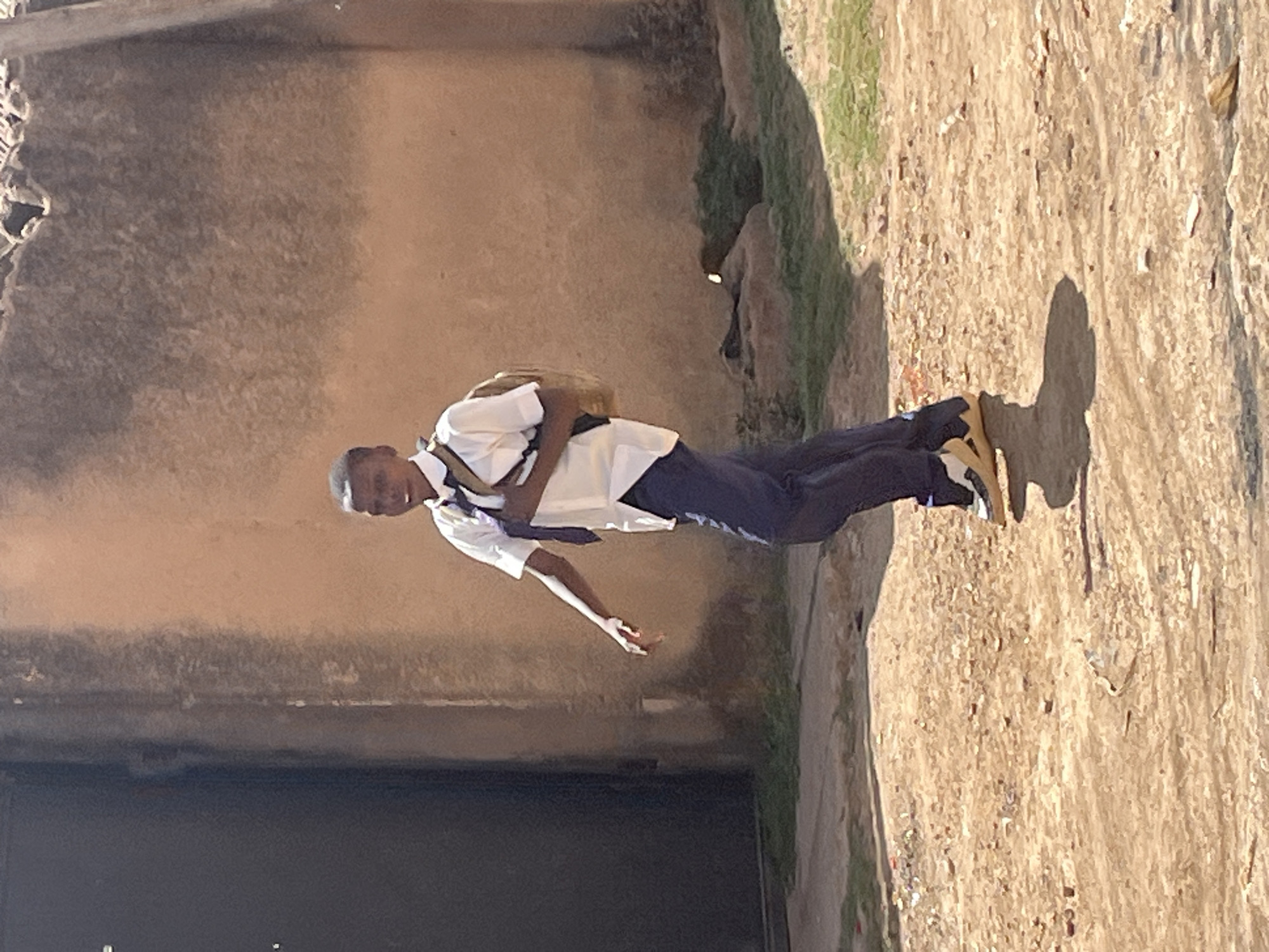 A young African man in a school uniform walks on the side of a street in Barakin Akawo, Nigeria. He wears a white shirt, dark pants, and brown shoes. He has a backpack on his back. The street is unpaved, with a wall on one side and a grassy area on the other. The sun is shining, casting a shadow of the man on the ground.