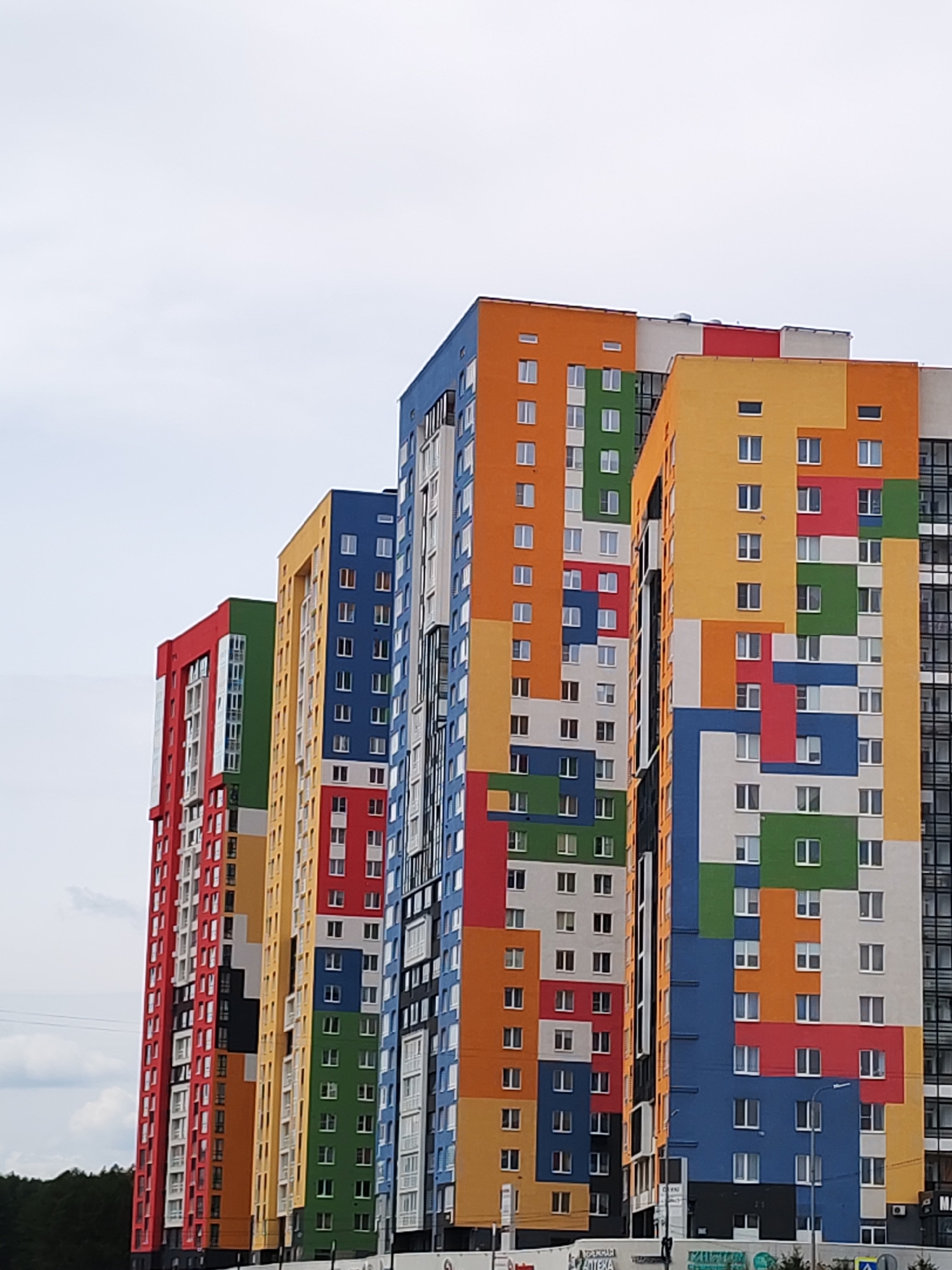 The image shows four tall residential buildings with colorful facades. The buildings are located in the city of Izhevsk, Russia. Each building has a distinct color scheme: red, blue, orange, green, and yellow. The facades feature a geometric pattern with rectangular shapes in various colors. The buildings are tall and multi-storied with numerous windows. The sky is overcast with clouds. The base of the buildings are obscured by trees and signage.
