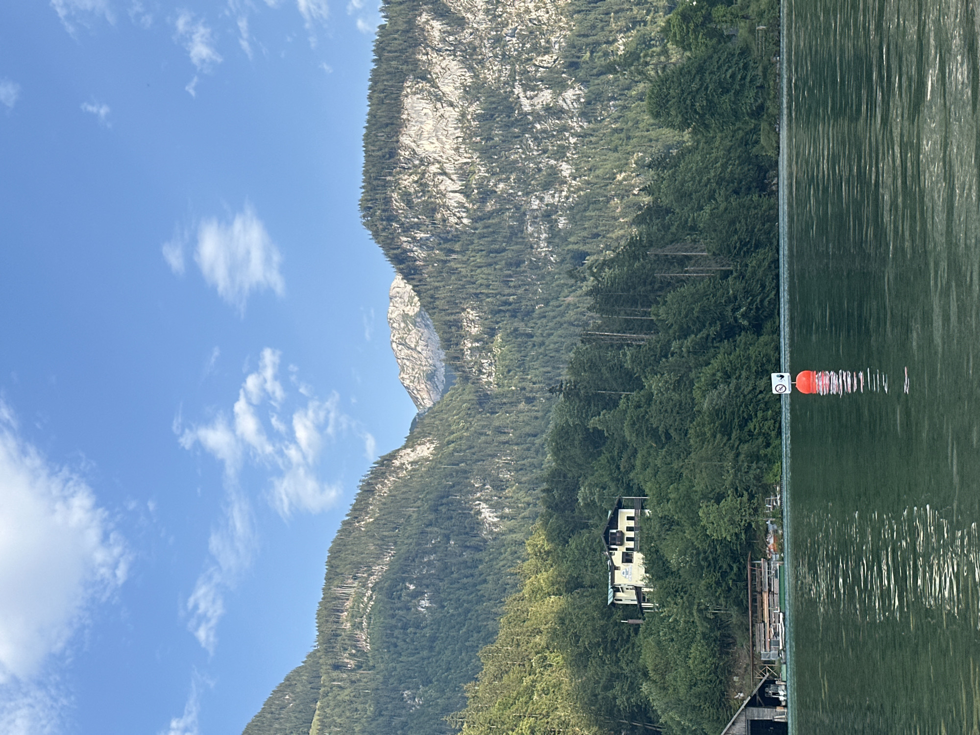 The image shows a serene lake with a mountain backdrop in Schönau am Königssee, Germany. The water in the foreground is a deep green, reflecting the surrounding trees and sky. A bright orange buoy with a sign depicting a no-entry symbol is visible in the middle of the lake. On the left side of the frame, a wooden boathouse with several boats docked is nestled amongst lush green trees.
A yellow house with a dark roof stands prominently on a hillside overlooking the lake, surrounded by dense evergreen forests. The mountains behind the house are covered in trees, with some rocky, exposed faces visible towards the peaks. The sky is a clear blue with scattered white clouds, suggesting a pleasant day. The overall scene evokes a sense of tranquility and natural beauty.