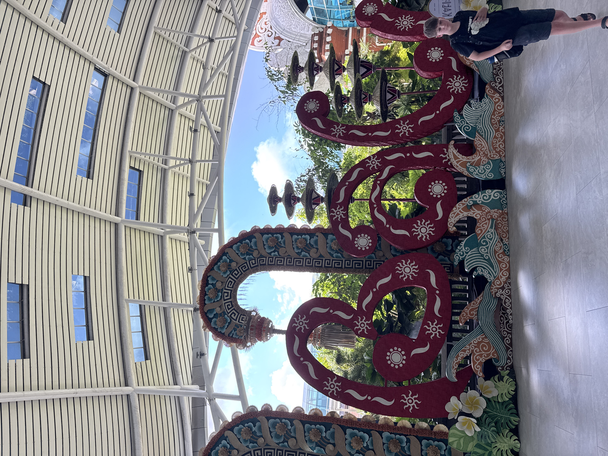 The image captures a large, decorative installation spelling out "BALI" in bright red, stylized letters. The letters are adorned with sun motifs and wave-like patterns at their base, with tropical foliage and flowers flanking the word. Behind the installation, a covered walkway with a sloped, ribbed ceiling and skylights is visible, offering a glimpse of a bright blue sky with scattered clouds. The setting appears to be an indoor or semi-outdoor public space, possibly an airport or a cultural center, given the architectural style and the presence of lush greenery.
To the right of the "BALI" installation, a young boy wearing a black t-shirt and shorts walks past, carrying a backpack. He is the only person prominently featured in the scene. In the background, behind the foliage, glimpses of a building with traditional Indonesian architectural elements can be seen, including ornate carvings and a red-tiled roof, suggesting a location in Bali, Indonesia.
While the prompt specifies the city of Kuta, Indonesia, there are no direct visual cues in the image that explicitly confirm this location. However, the presence of the "BALI" signage and the architectural style are consistent with a location in Bali. The overall scene conveys a festive and welcoming atmosphere, celebrating the identity of the island.