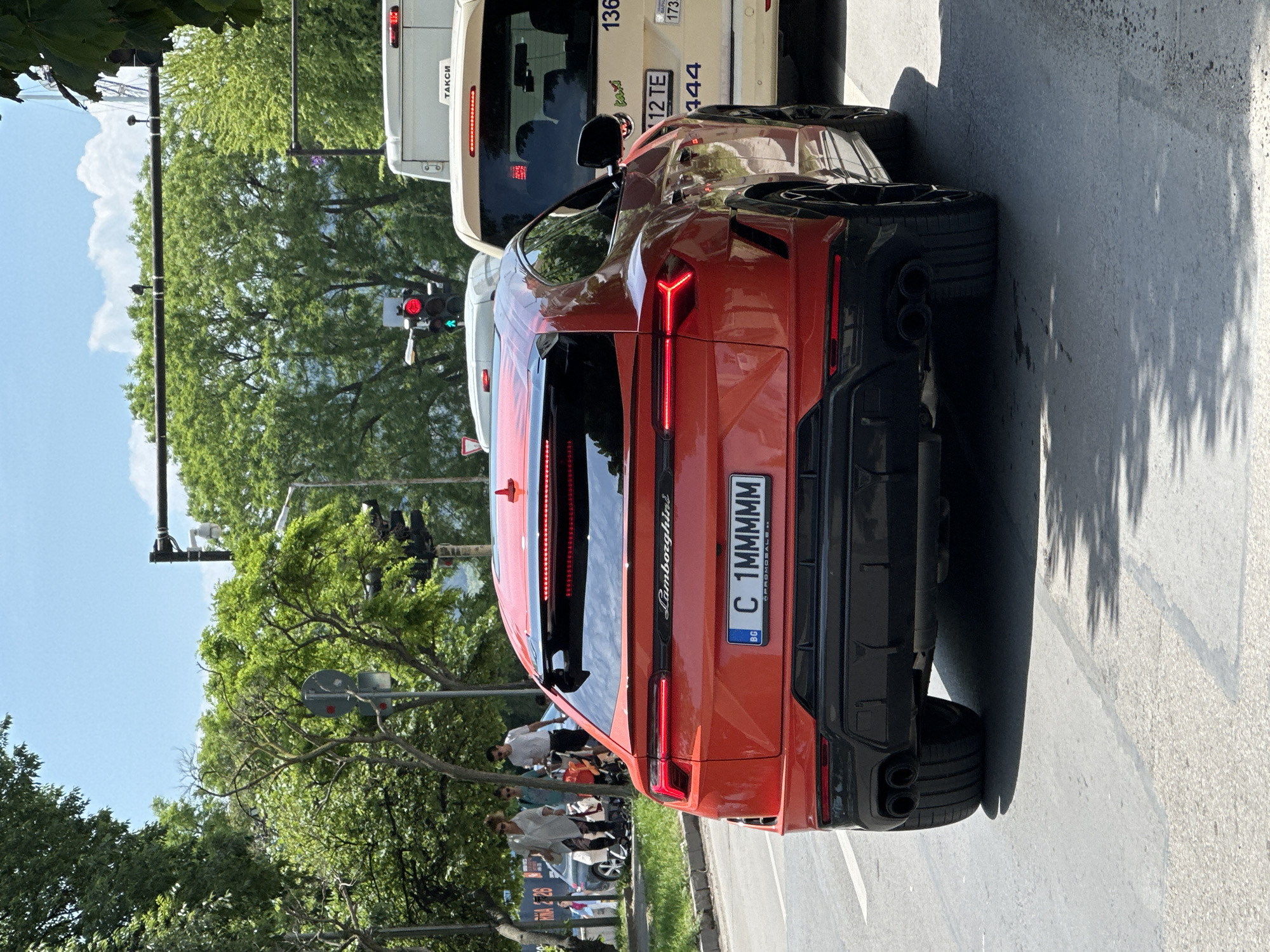 The image captures the rear of an orange Lamborghini Urus stopped on a street in Varna, Bulgaria. The car has a Bulgarian license plate "C 1MMMMM." A white taxi is partially visible behind the Lamborghini. People are walking on the sidewalk to the left of the car, near trees and vegetation. A traffic light is seen in the background. The weather appears to be sunny.