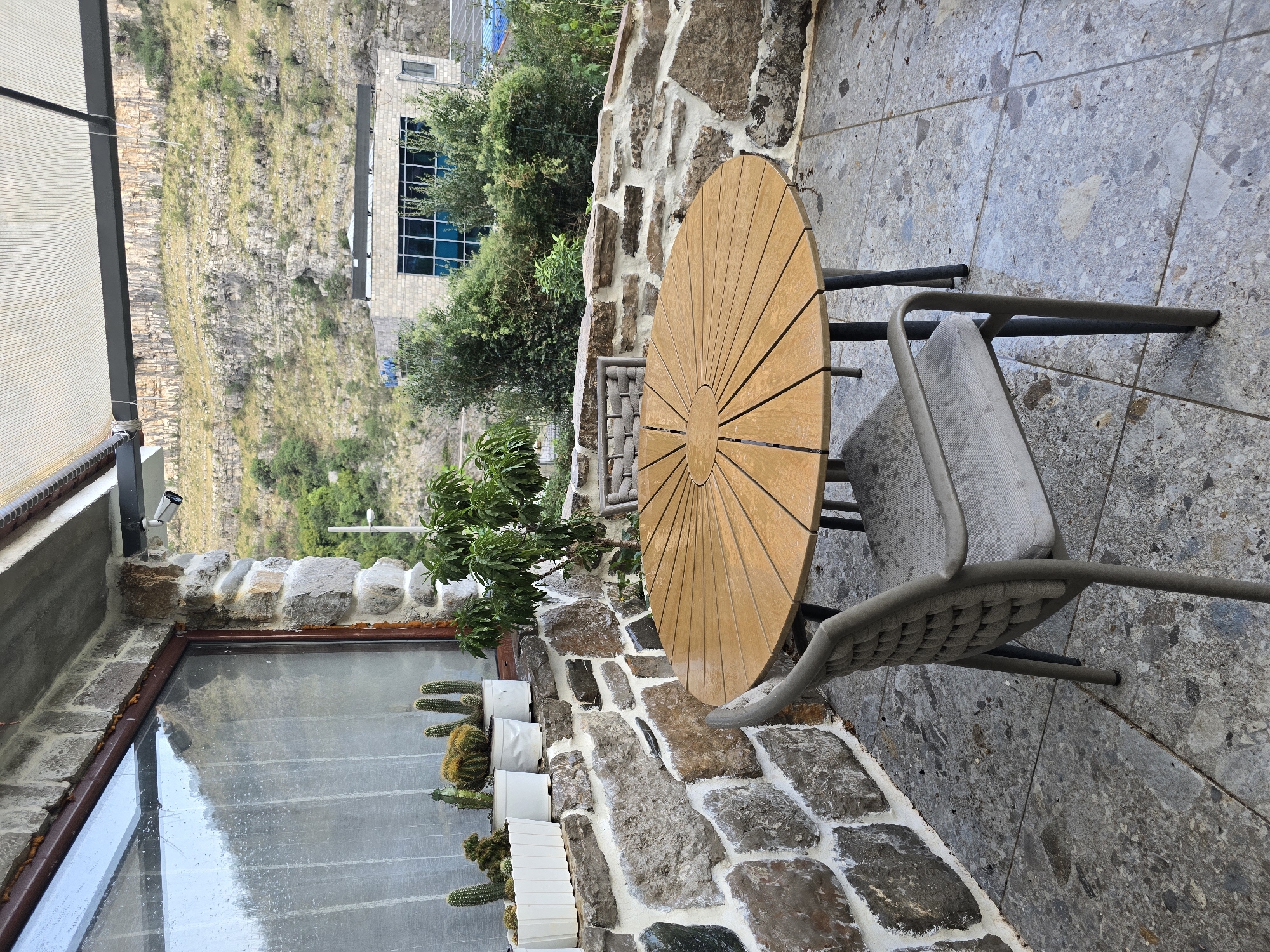 The image depicts an outdoor patio area with a round wooden table and two chairs. The patio floor is paved with large, gray, speckled tiles. Behind the table and chairs is a stone wall, which forms the base of a covered structure. The wall has a rough, natural appearance, with large stones of varying shapes and colors mortared together. Above the wall, a window with a condensation-covered pane looks out onto a natural landscape. Potted cacti are arranged on a ledge along the top of the stone wall beneath the window. To the right of the table, a small tree with green leaves is visible.
Beyond the patio, the background reveals a steep, rocky hillside dotted with vegetation. In the middle ground, a modern building with a light-colored stone facade and large blue-tinted windows is situated against the hillside. The environment appears to be somewhat remote, with a focus on natural elements and a contrast between the rustic stone architecture and the modern building. The overall scene suggests a peaceful, possibly rural or semi-rural, setting.
While the image does not explicitly show any people or actions, the presence of the table and chairs implies a space designed for outdoor dining or relaxation. The weather appears to be overcast or perhaps recently rained, given the condensation on the window and the damp appearance of the patio tiles. The location is described as Podgorica, Montenegro.