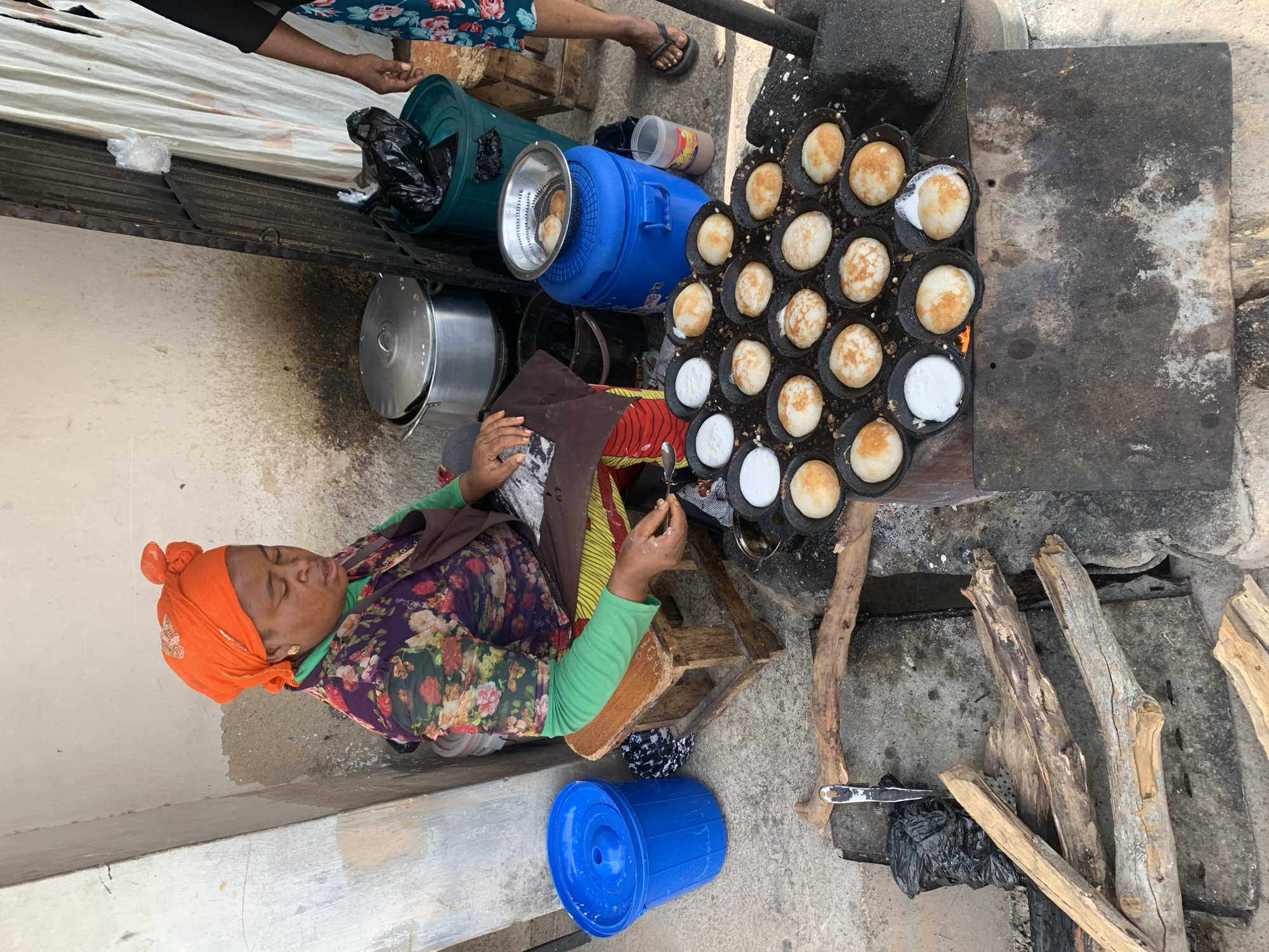 A woman in Jos, Nigeria, sits on a small wooden stool next to a charcoal stove, cooking small, round cakes in a multi-cupped pan. She wears an orange headscarf, a floral shirt, and green sleeves. Various blue and green containers are around her, along with firewood on the ground. A metal pot sits behind her, and a glimpse of another person is visible above the containers.