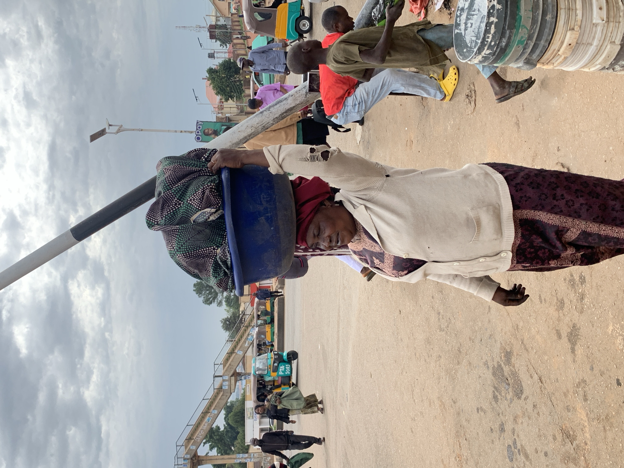 In Gura Top, Nigeria, a woman walks carrying a large blue basin filled with patterned fabric on her head. She wears a maroon headwrap, a cream-colored cardigan, and a dark patterned skirt. In the background, a pedestrian bridge crosses over a street. Several people are visible, including men and women walking, and children. A bright teal and yellow motorized tricycle, a common mode of transport in the region, is parked nearby, along with other vehicles. The ground is a dusty, paved surface, and the sky is overcast with scattered clouds.